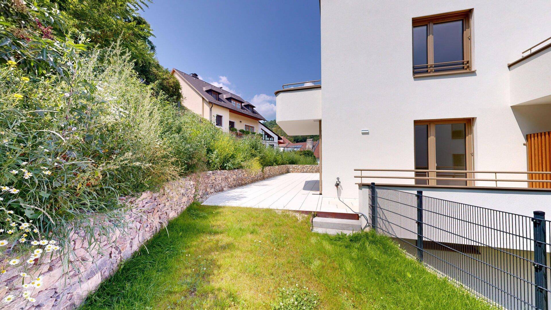 Grüner Gartenbereich mit Rasenfläche und gepflasterter Terrasse neben dem modernen Gebäude.