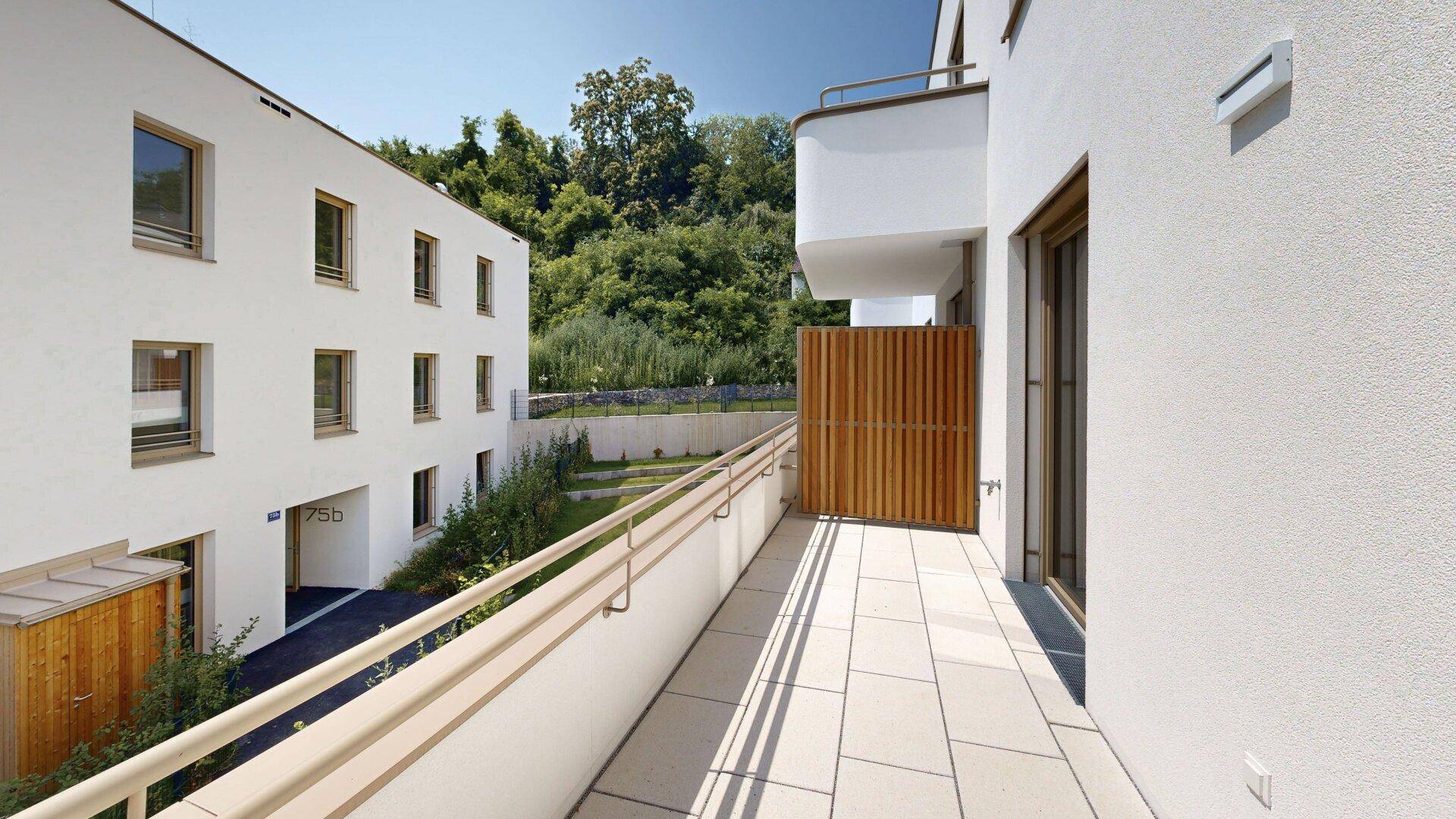 Langer Balkon mit Fliesenboden und Holzelementen, umgeben von modernen Gebäuden und viel Grün.