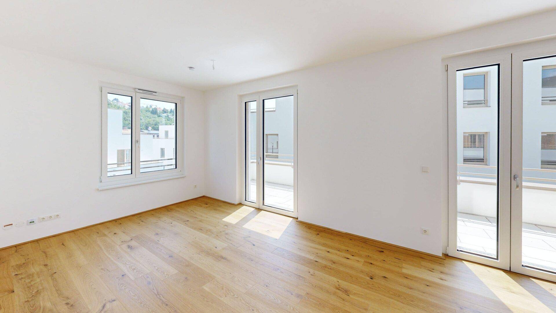 Helles, leeres Zimmer mit Holzboden und großen Fenstern, die Zugang zu einem Balkon bieten.