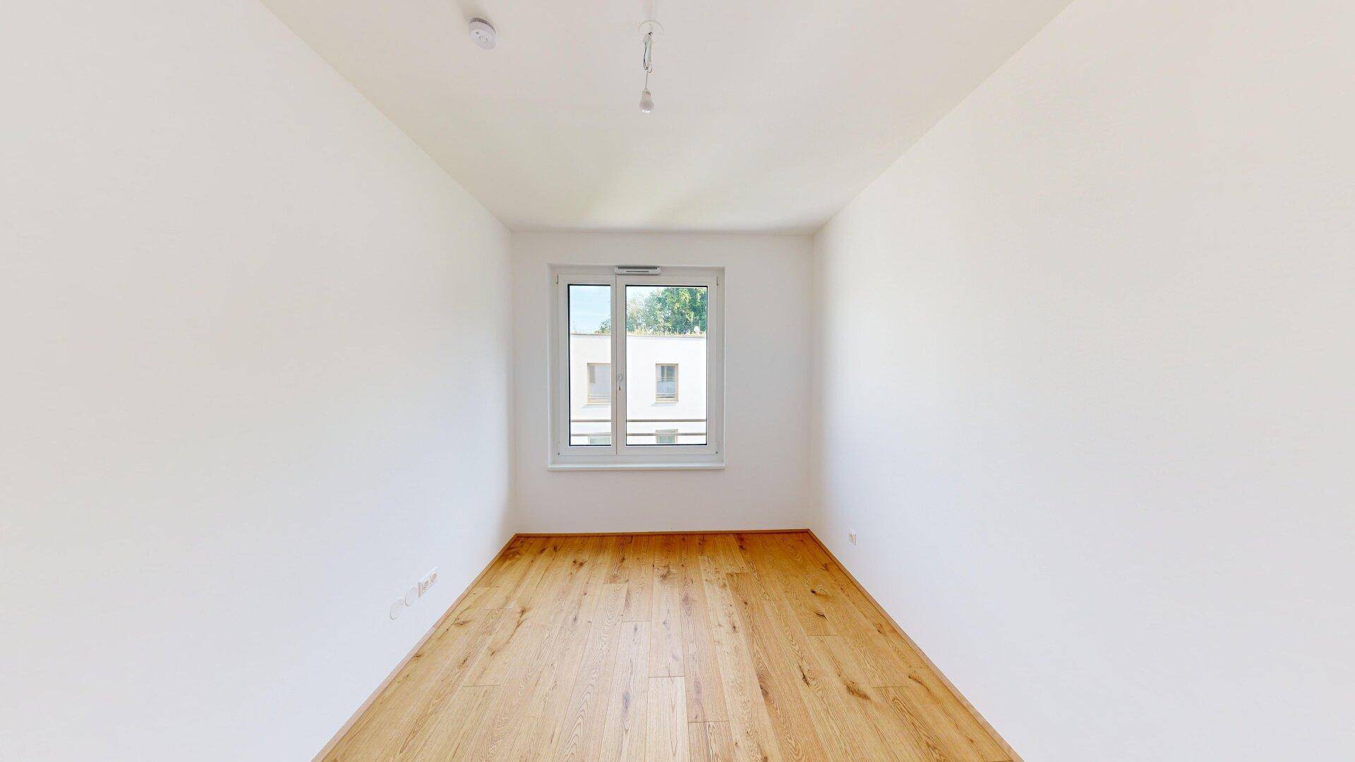 Leeres Schlafzimmer mit hellem Parkettboden, weißen Wänden und einem großen Fenster für Tageslicht.