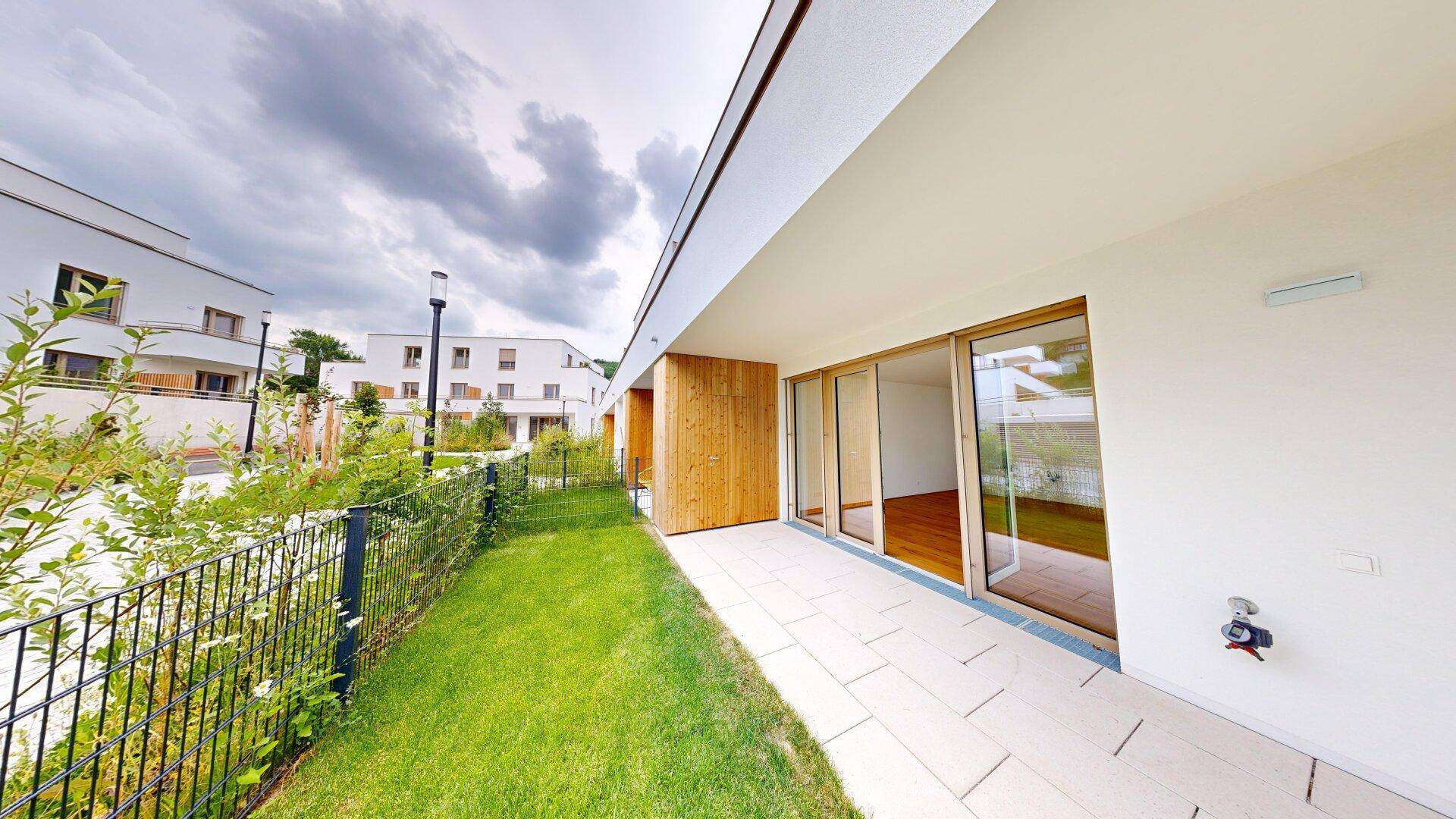 Moderne Terrasse mit Fliesenboden und Zugang zum Garten, umgeben von grüner Landschaft und neuen Gebäuden.