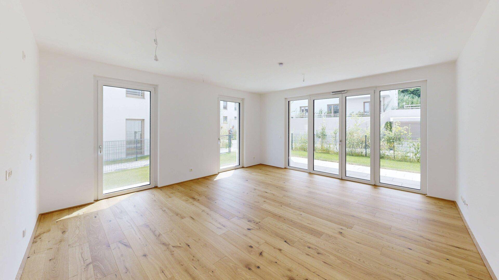 Heller, leerer Wohnraum mit großen Fenstern und Holzboden, bietet Zugang zum Außenbereich.