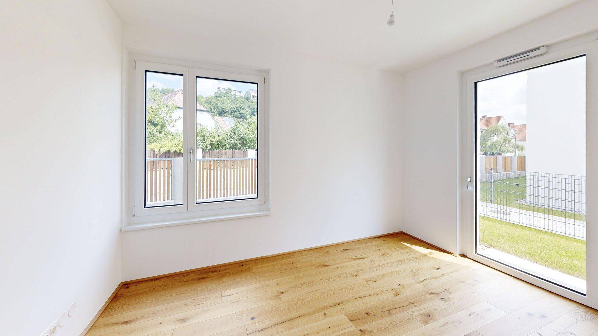 Helles Zimmer mit Holzboden und Fenster, das einen Blick ins Grüne bietet und Zugang nach draußen hat.