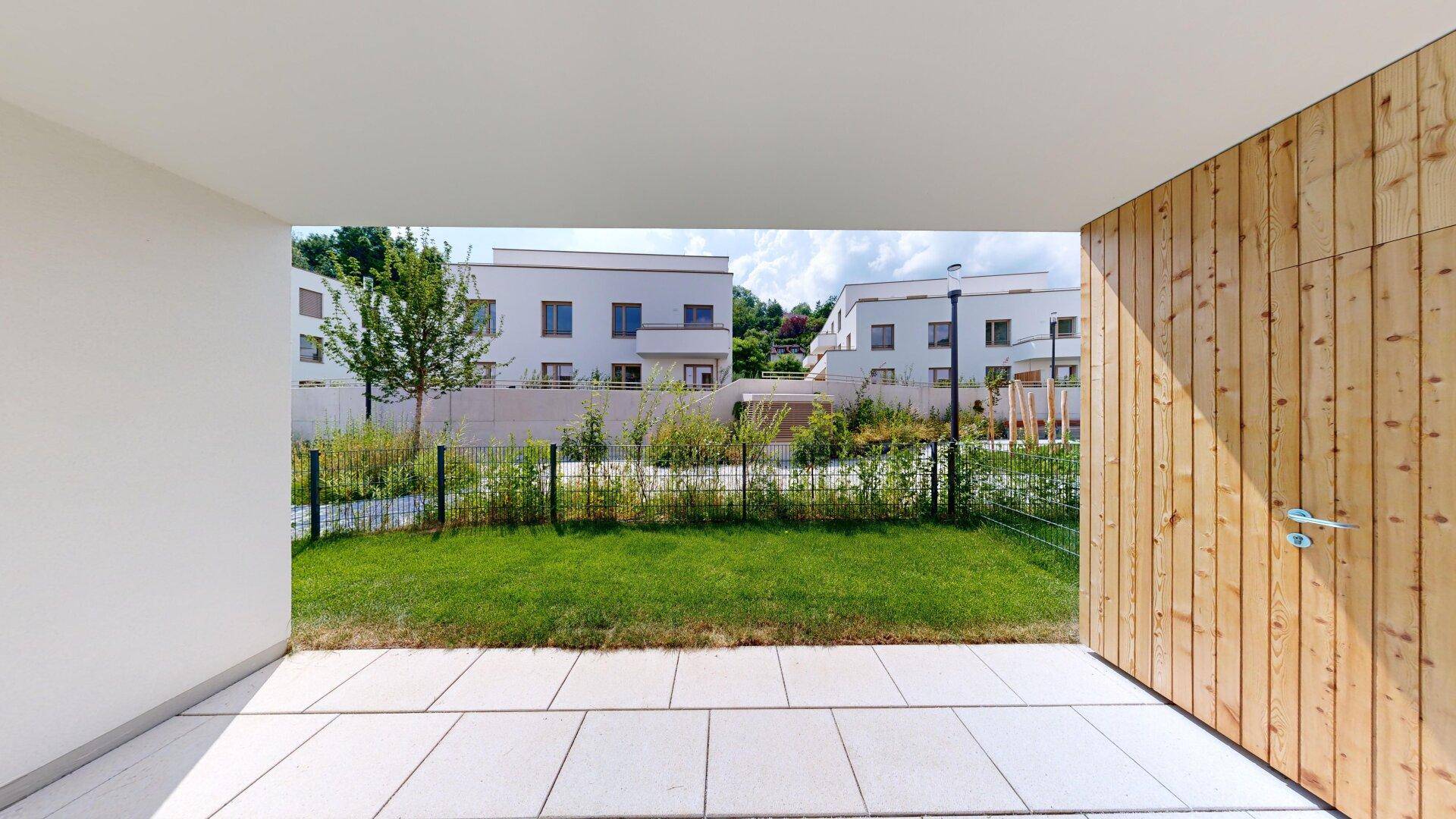 Blick von der überdachten Terrasse auf den kleinen Garten und die umliegenden modernen Gebäude.