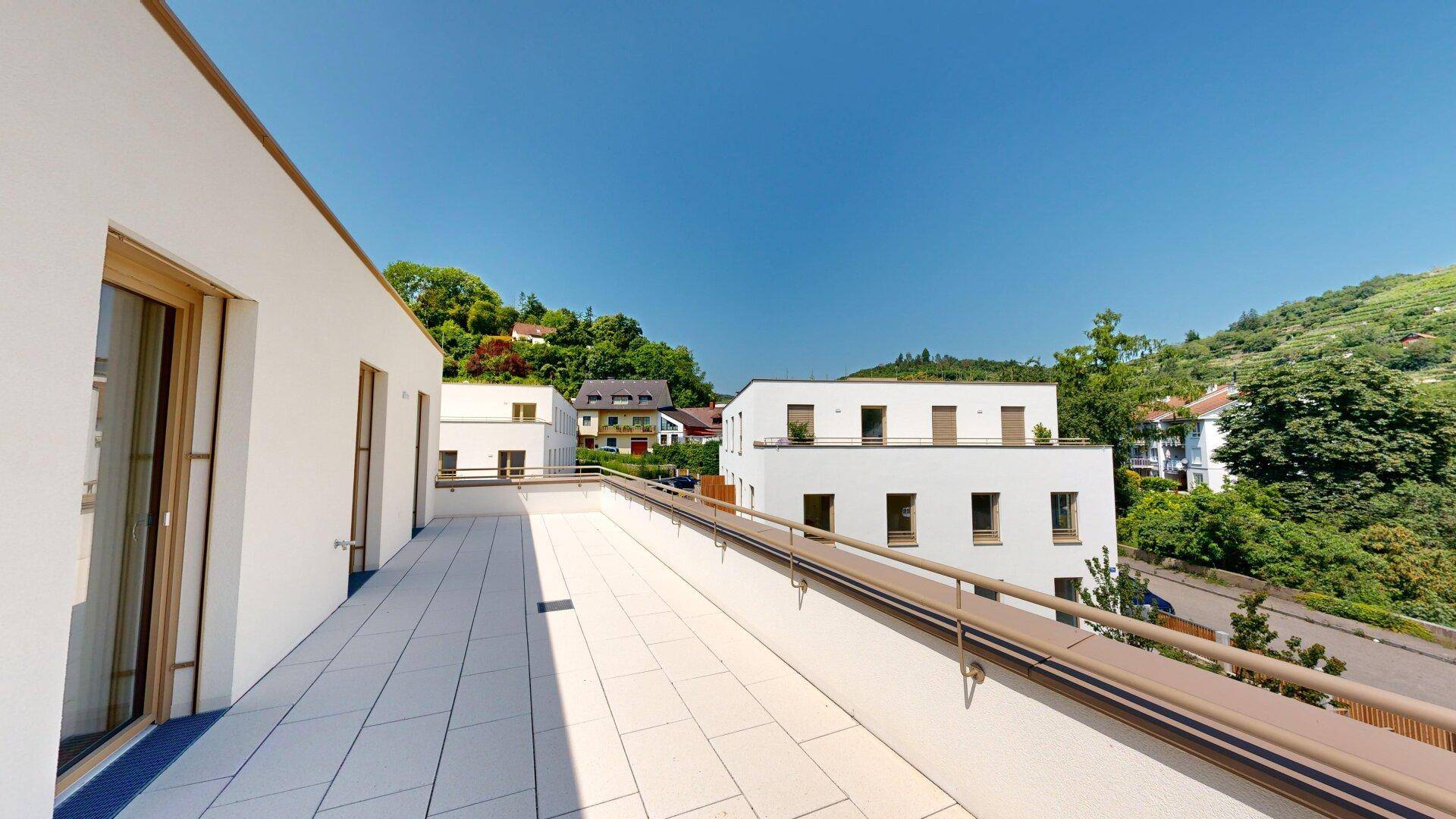 Große, sonnige Terrasse mit Fliesenboden und Blick auf die umliegende Landschaft und Gebäude.