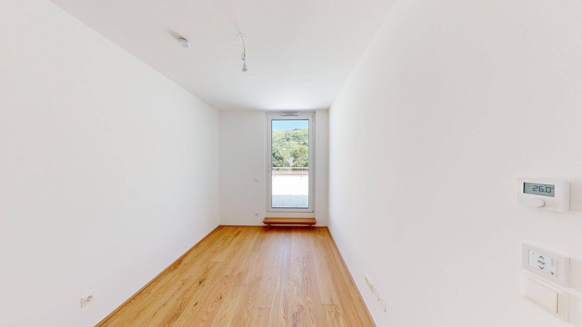 Helles Zimmer mit Holzboden und Zugang zu einem Balkon oder einer Terrasse, mit Temperaturregler.