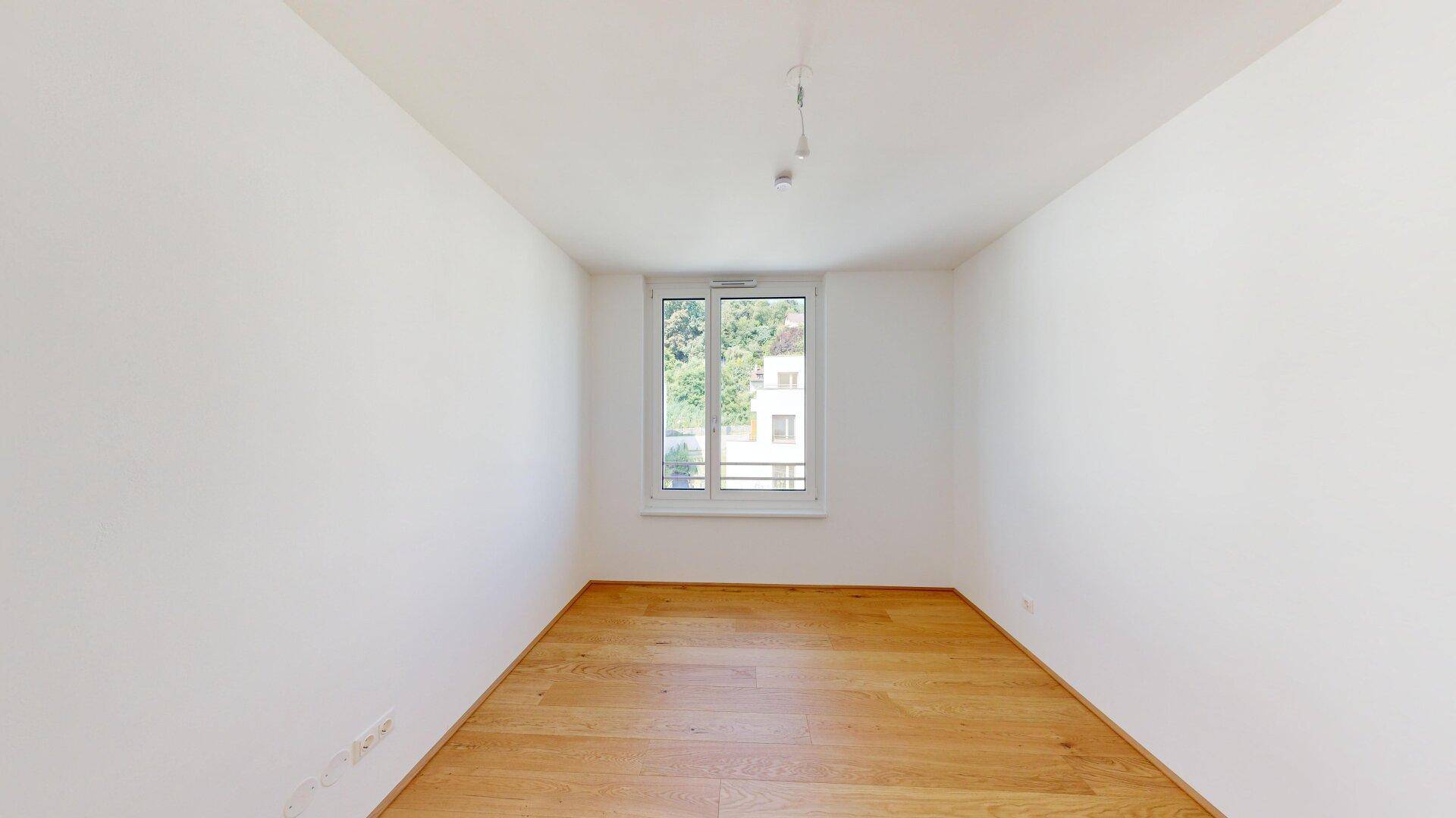 Ruhiges Schlafzimmer mit hellem Parkettboden und einem Fenster mit Blick ins Grüne.