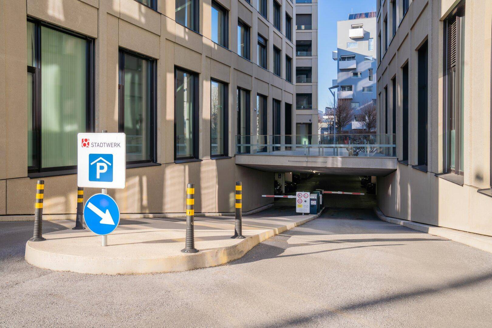 Zufahrt zu einer Tiefgarage mit Schranke und Beschilderung, umgeben von moderner Gebäudearchitektur.