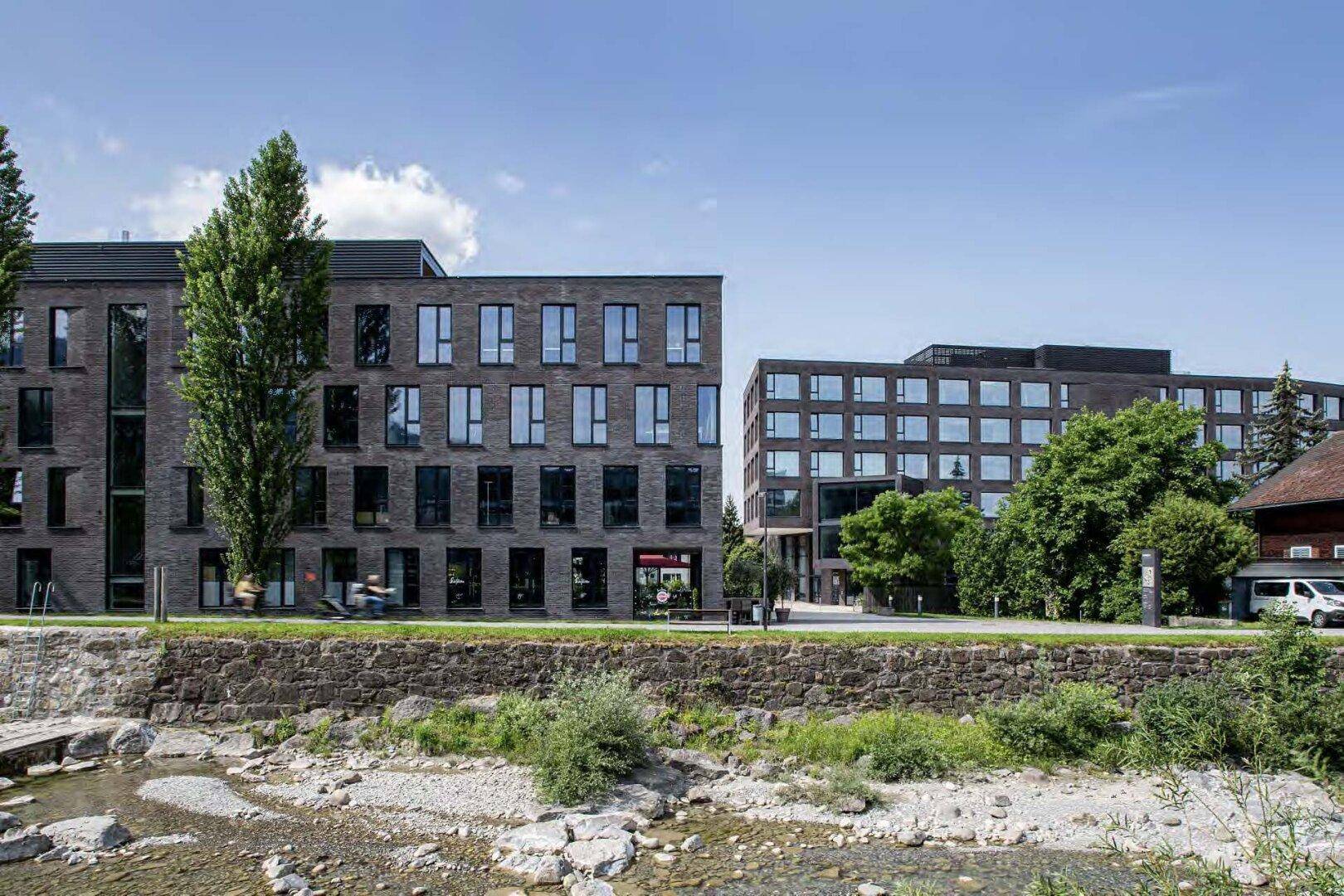 Moderne Bürogebäude mit dunkler Ziegelfassade entlang eines kleinen Flusses unter blauem Himmel.