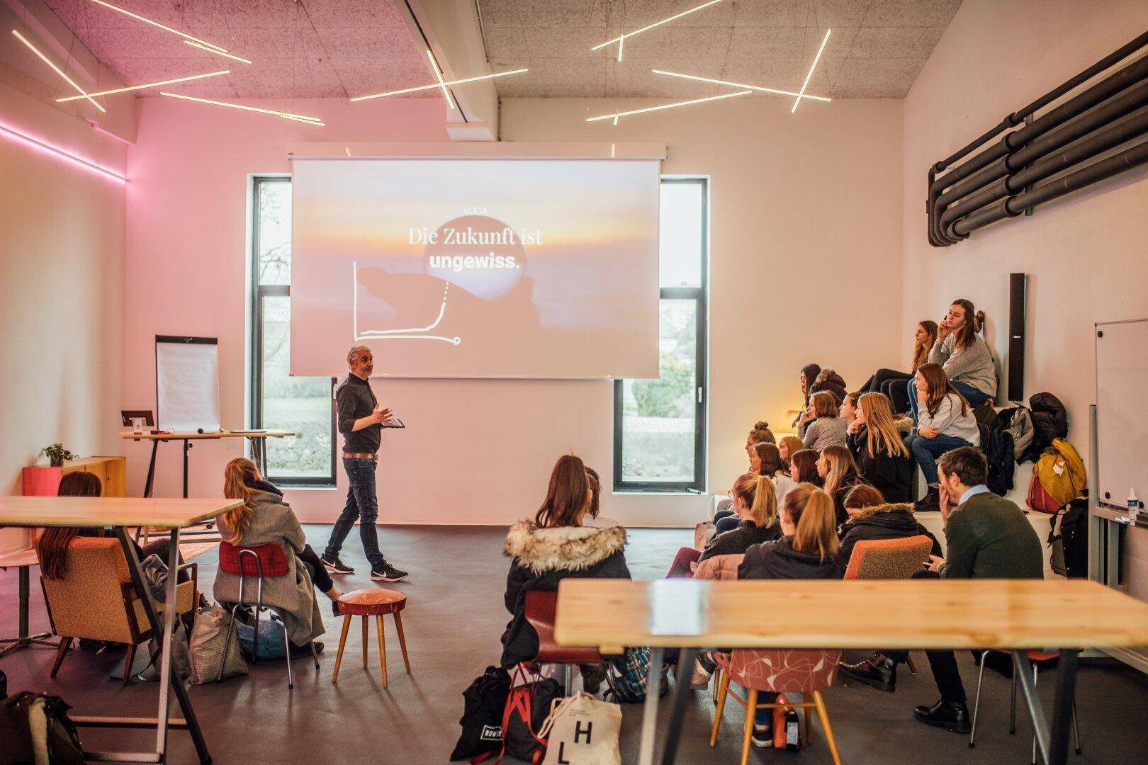 Moderner Konferenzraum mit Präsentationsleinwand, vielen Sitzgelegenheiten und kreativer Beleuchtung.