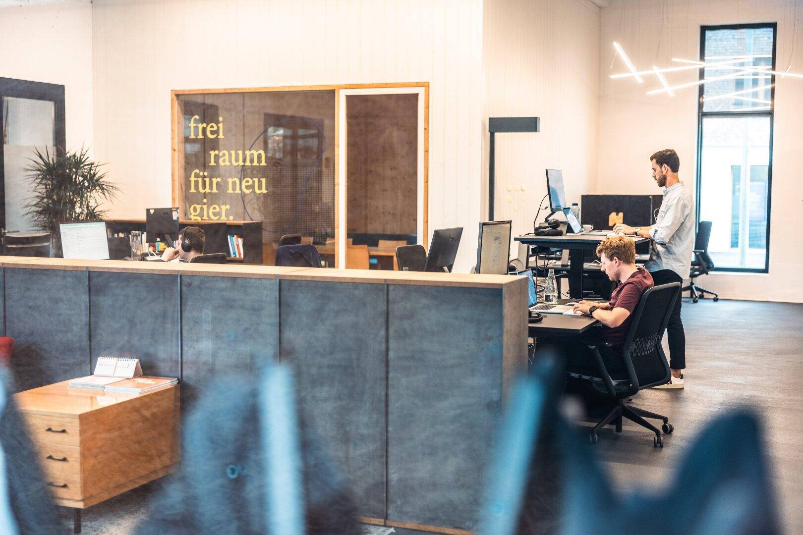 Helles Großraumbüro mit modernen Arbeitsplätzen, Stehtischen und kreativer Wandgestaltung.