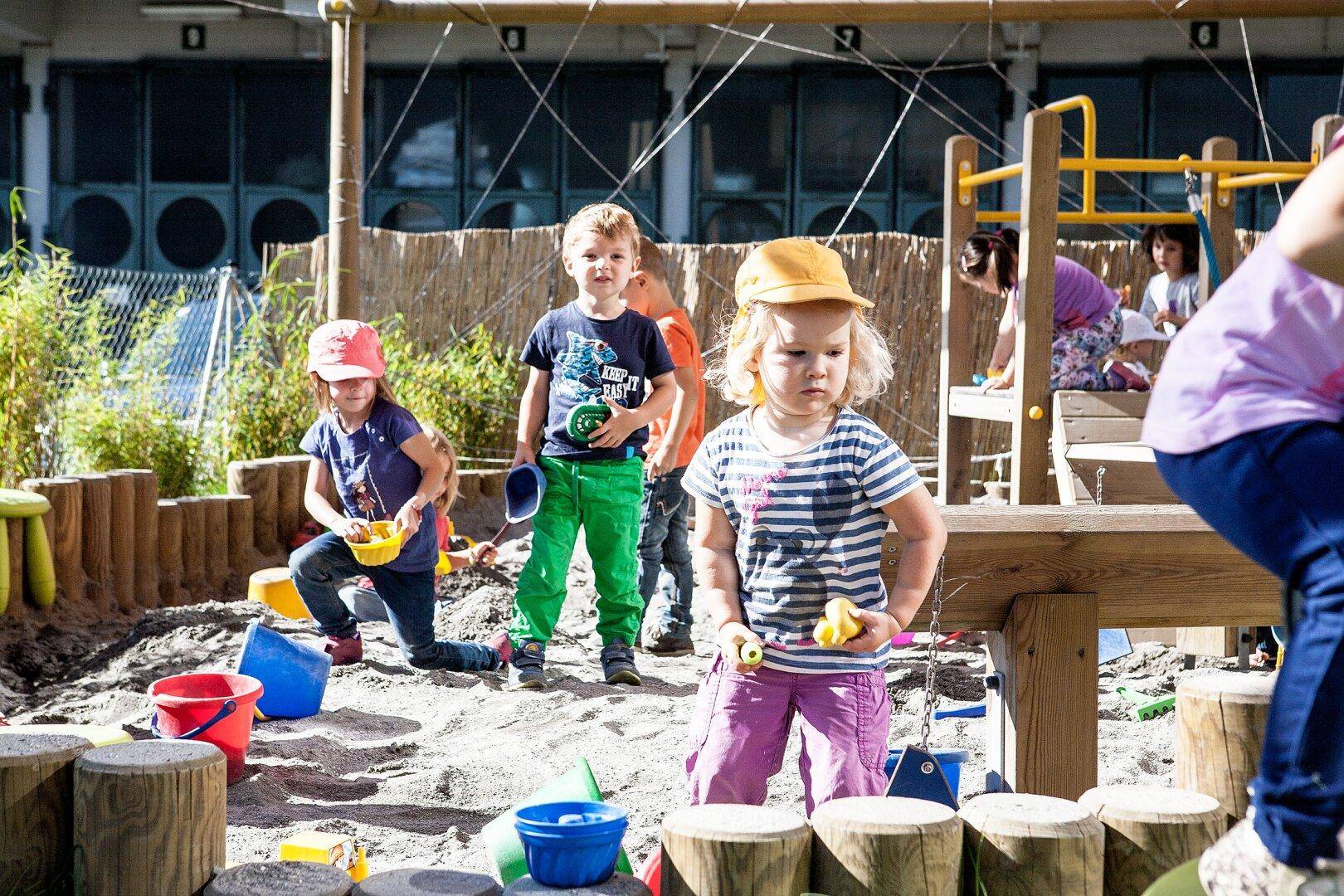 Kinder spielen in einem Sandkasten auf einem Spielplatz mit Holzelementen und grüner Umgebung.
