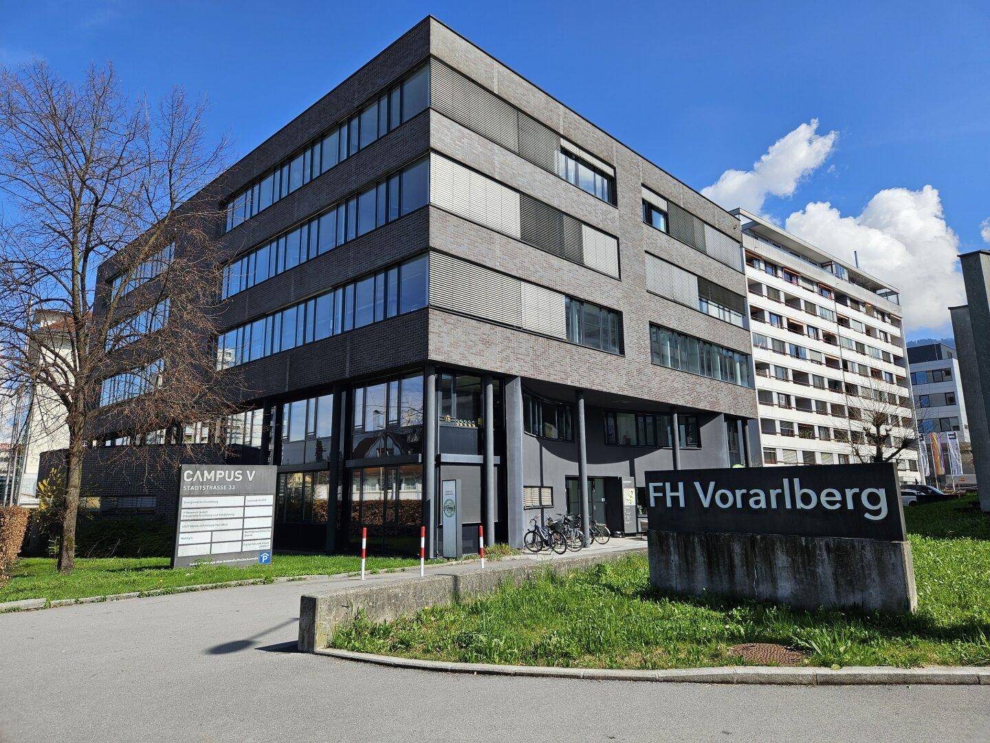 Eingangsbereich eines modernen Gebäudes mit FH Vorarlberg Beschilderung und Fahrradständern.