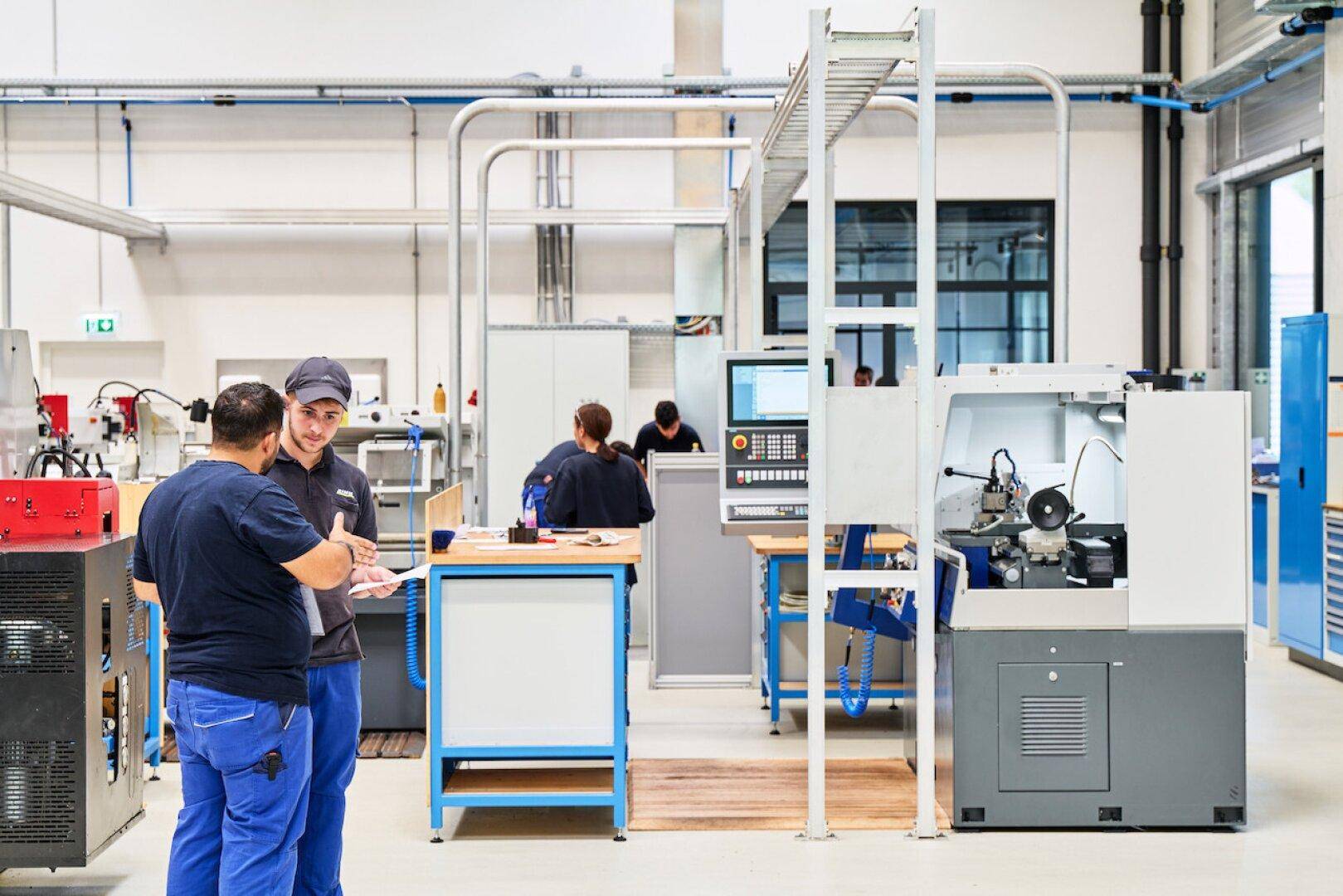 Mitarbeiter besprechen Details in einer modernen Werkstatt mit Präzisionsmaschinen und Arbeitsplätzen.