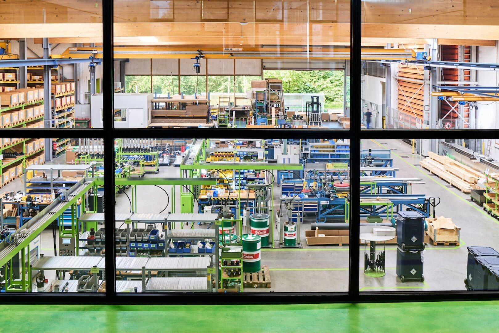 Blick durch ein großes Fenster in eine weitläufige Industriehalle mit Maschinen und Lagerbereichen.
