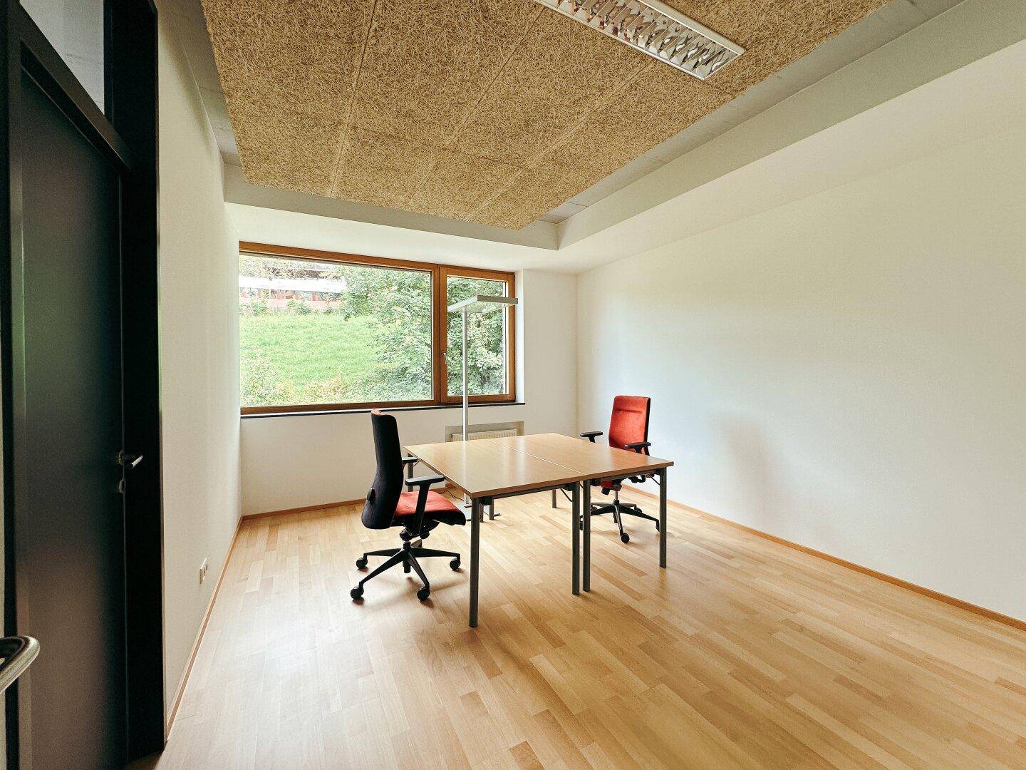 Helles Büro mit Holzboden, großem Fenster und Blick auf die umliegende Natur.