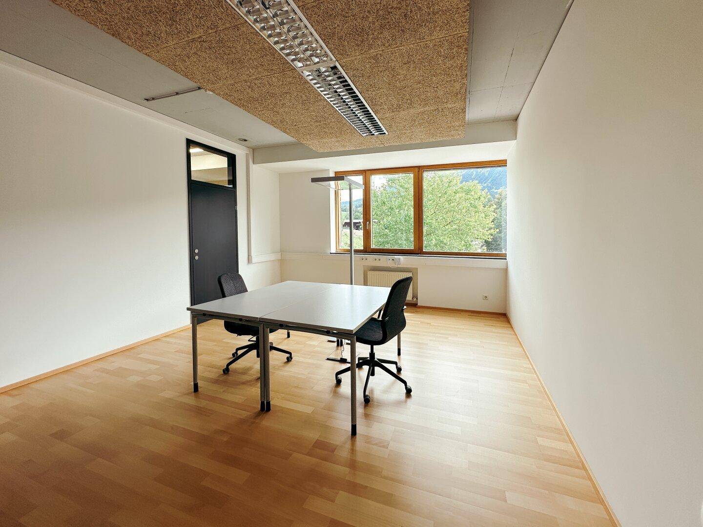 Heller Büroraum mit Holzboden, großen Fenstern und Heizkörper, bietet eine klare und funktionale Arbeitsfläche.