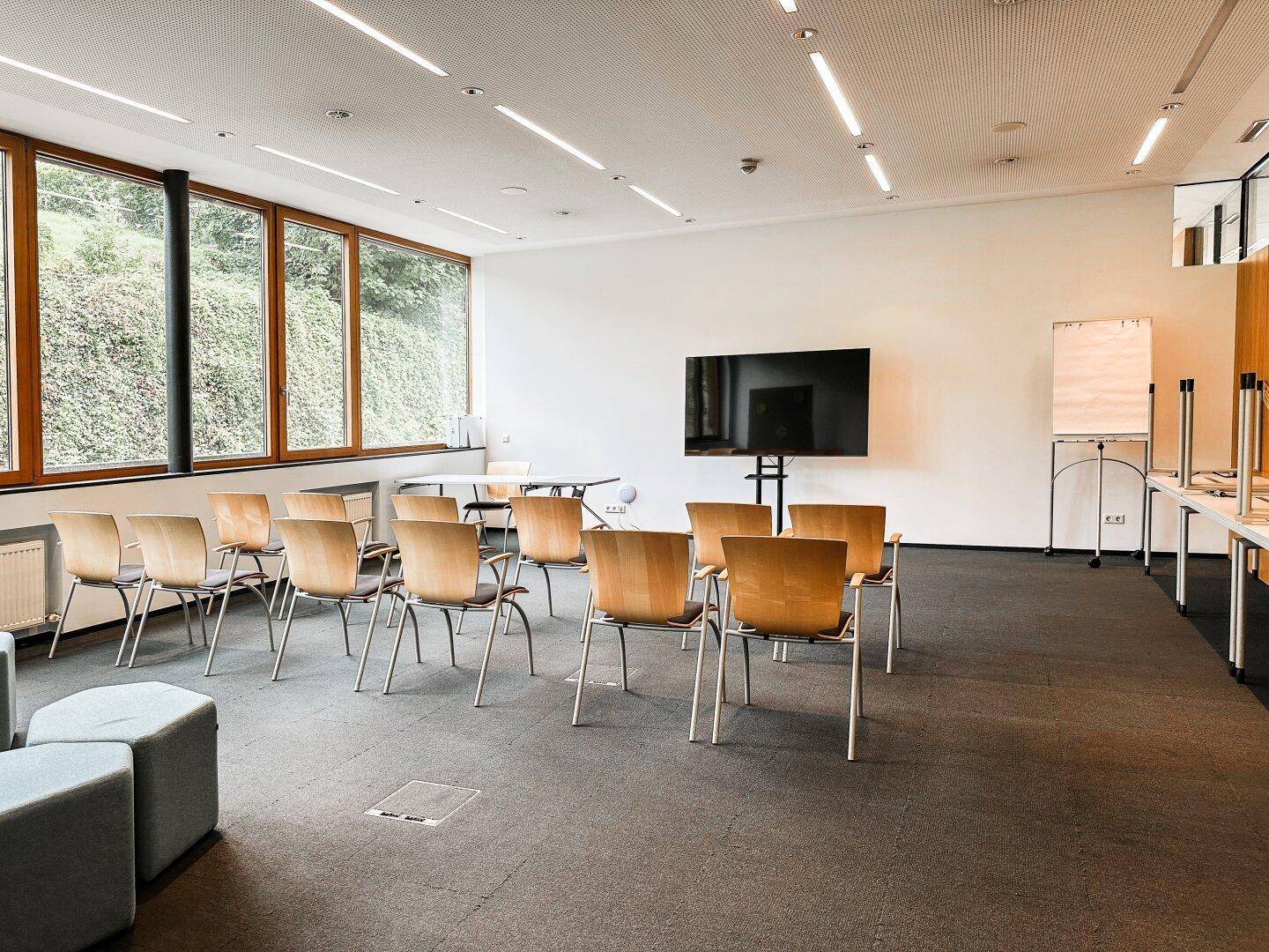 Großer Konferenzraum mit Reihen von Holzstühlen, einem Wandfernseher und großen Fenstern für Präsentationen.