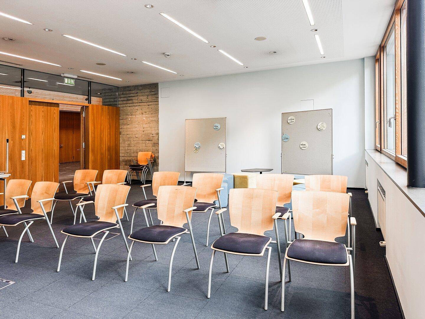 Großer Konferenzraum mit Reihen von Stühlen, Teppichboden und großen Fenstern für viel Tageslicht.