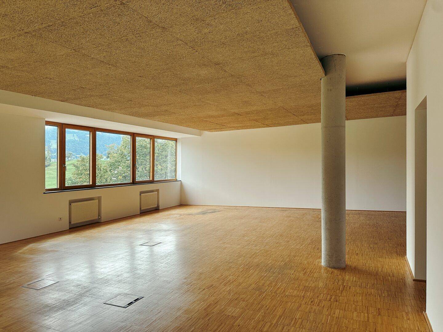 Geräumiger Innenraum mit Holzboden, Betonsäule und weitem Blick auf grüne Hügel und Berge.