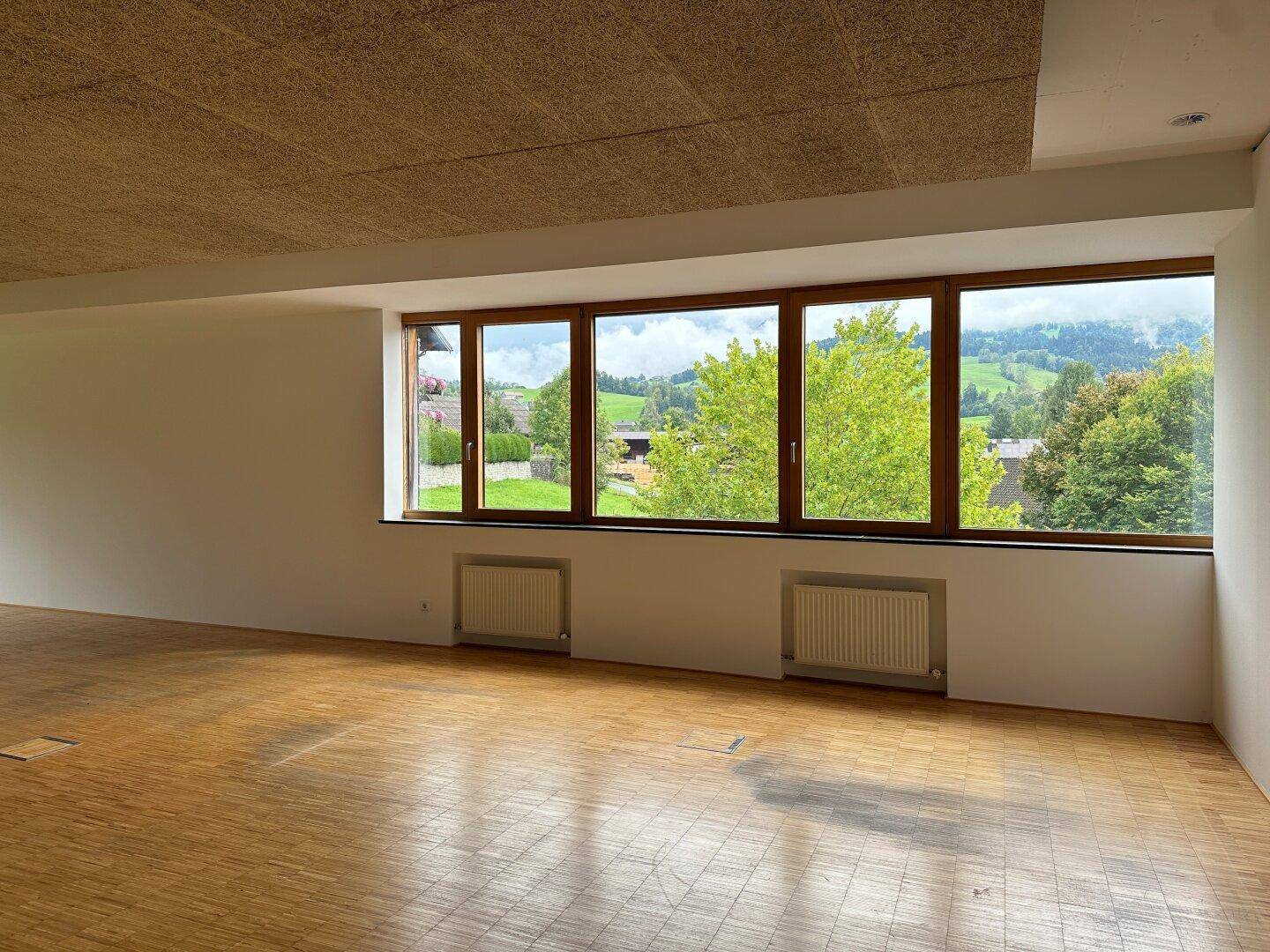 Große Holzfenster bieten einen malerischen Blick auf eine grüne Hügellandschaft mit vereinzelten Häusern.