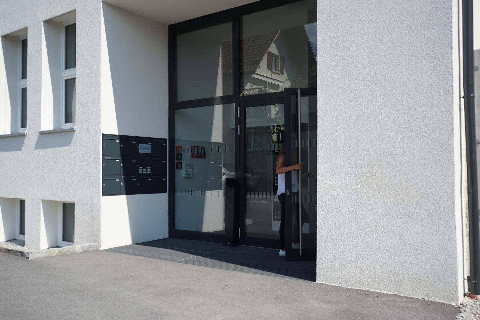 Eingangsbereich eines modernen Gebäudes mit großer Glastür und Briefkästen.