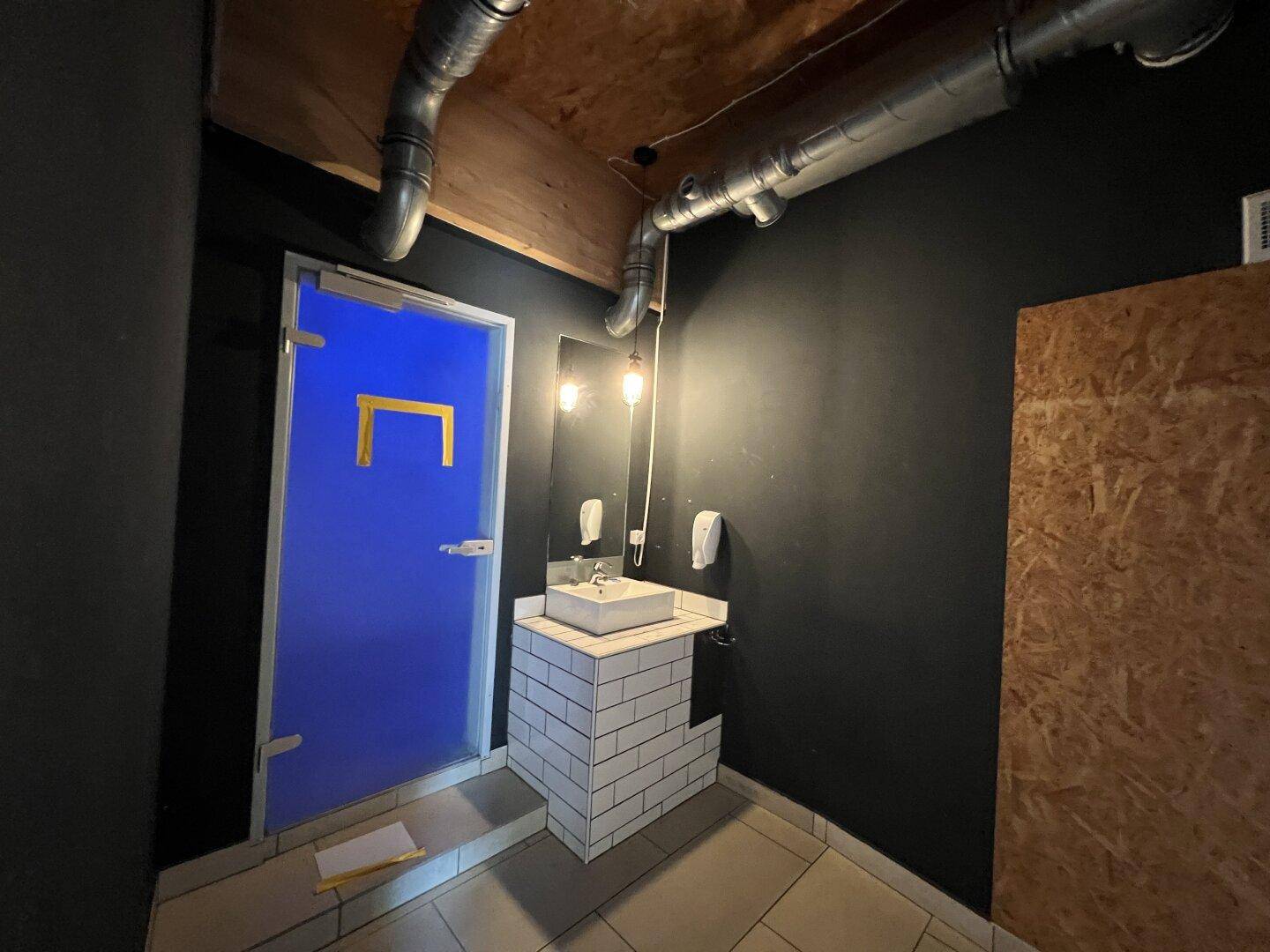 Kleine Toilette mit Waschbecken, blauer Tür und sichtbaren Lüftungsrohren an der Decke.