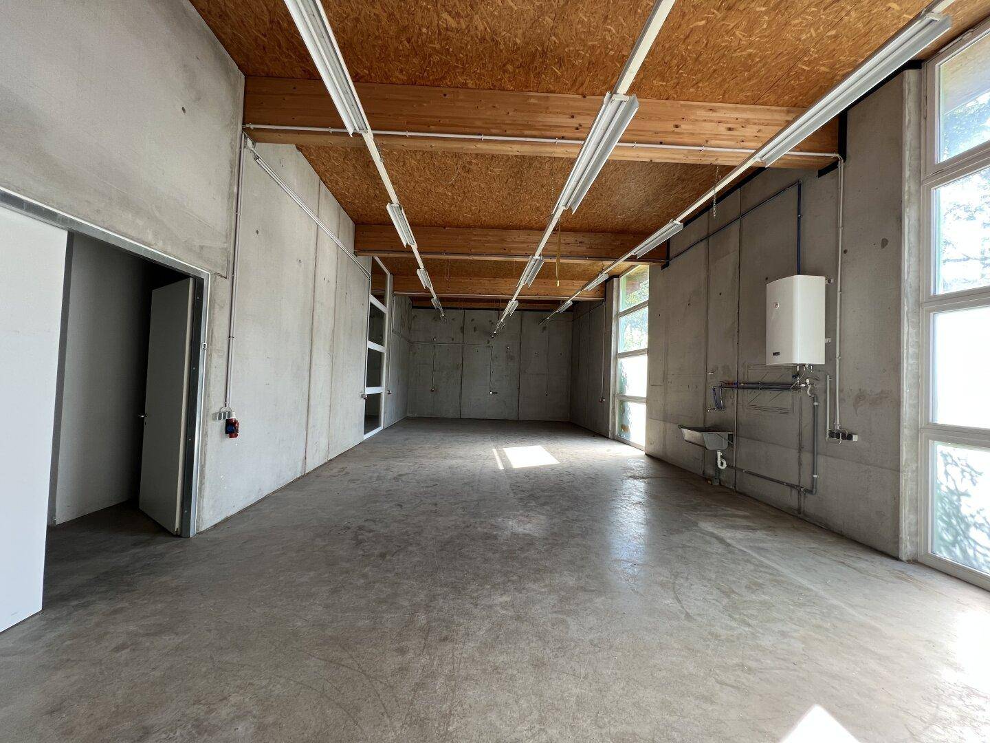 Geräumiger Industrieraum mit Betonwänden, hohen Fenstern und sichtbaren Installationen.