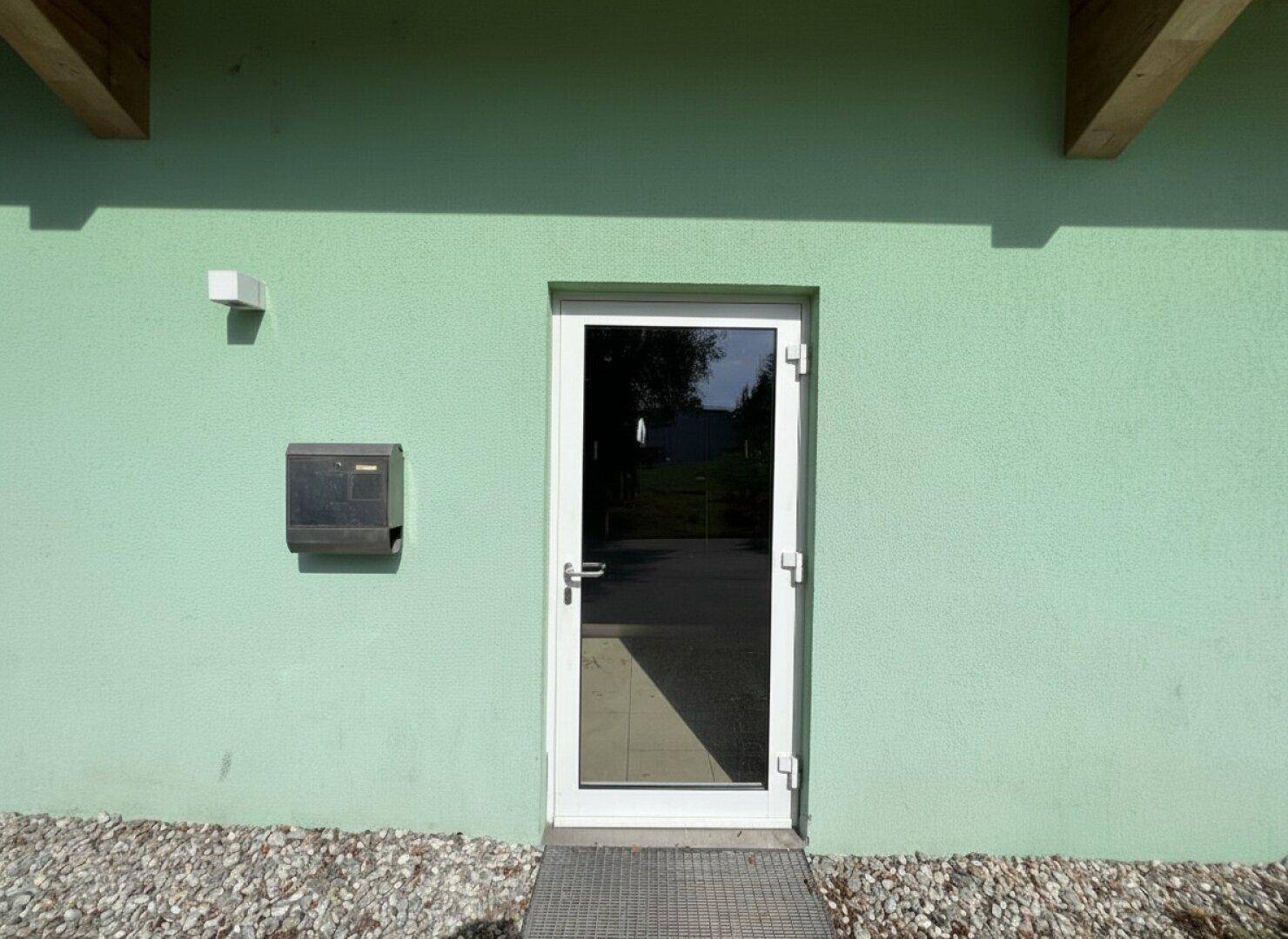 Eingangsbereich mit weißer Tür, grüner Fassade und Briefkasten an der Wand.