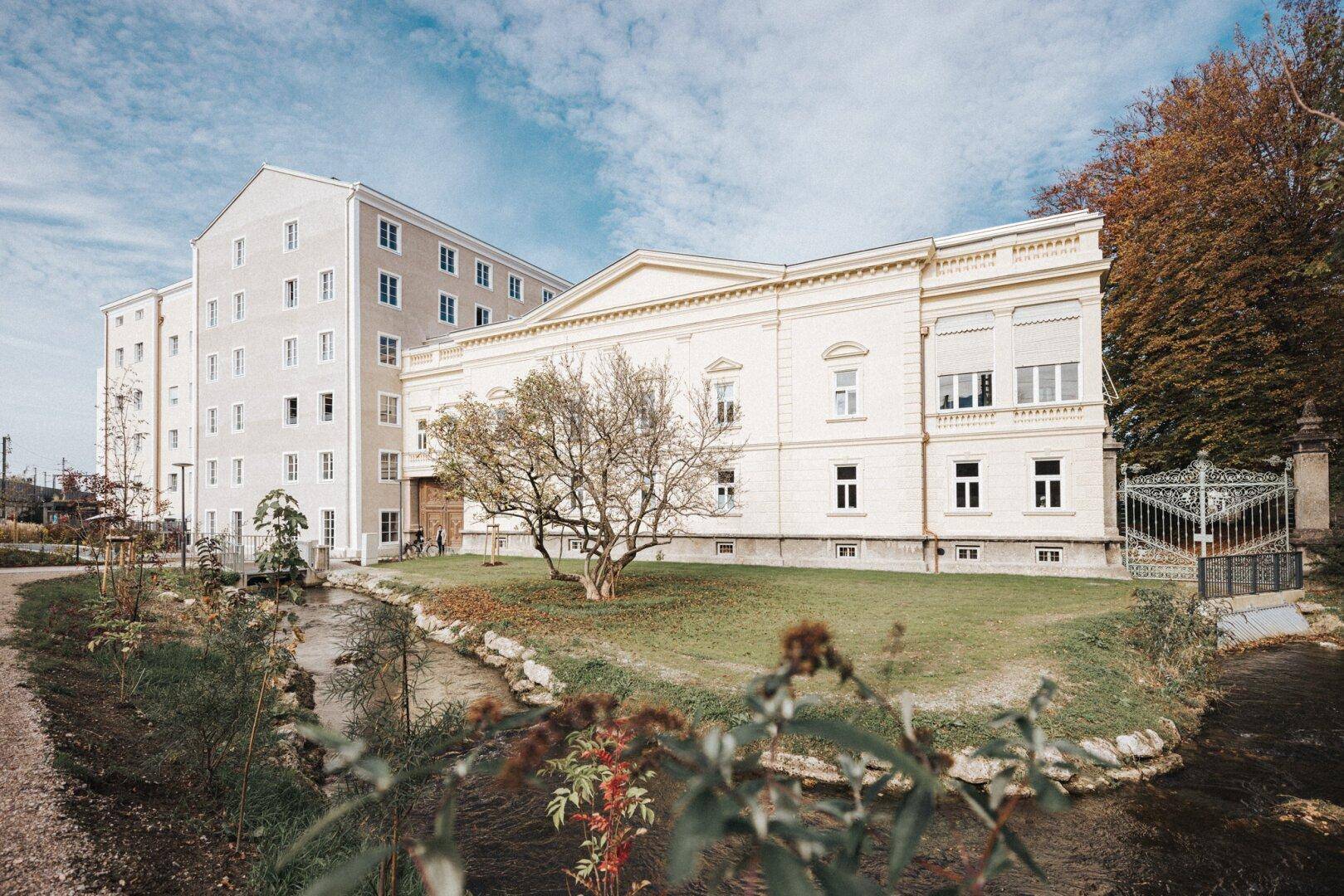 Historisches Gebäude mit klassischer Architektur neben einem modernen Anbau und einem Bachlauf.
