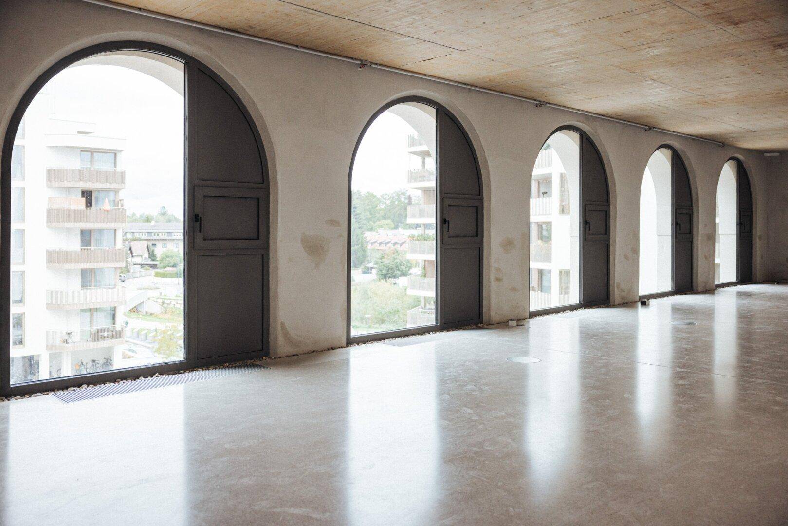 Großer, leerer Raum mit hohen Rundbogenfenstern und glänzendem Betonboden.