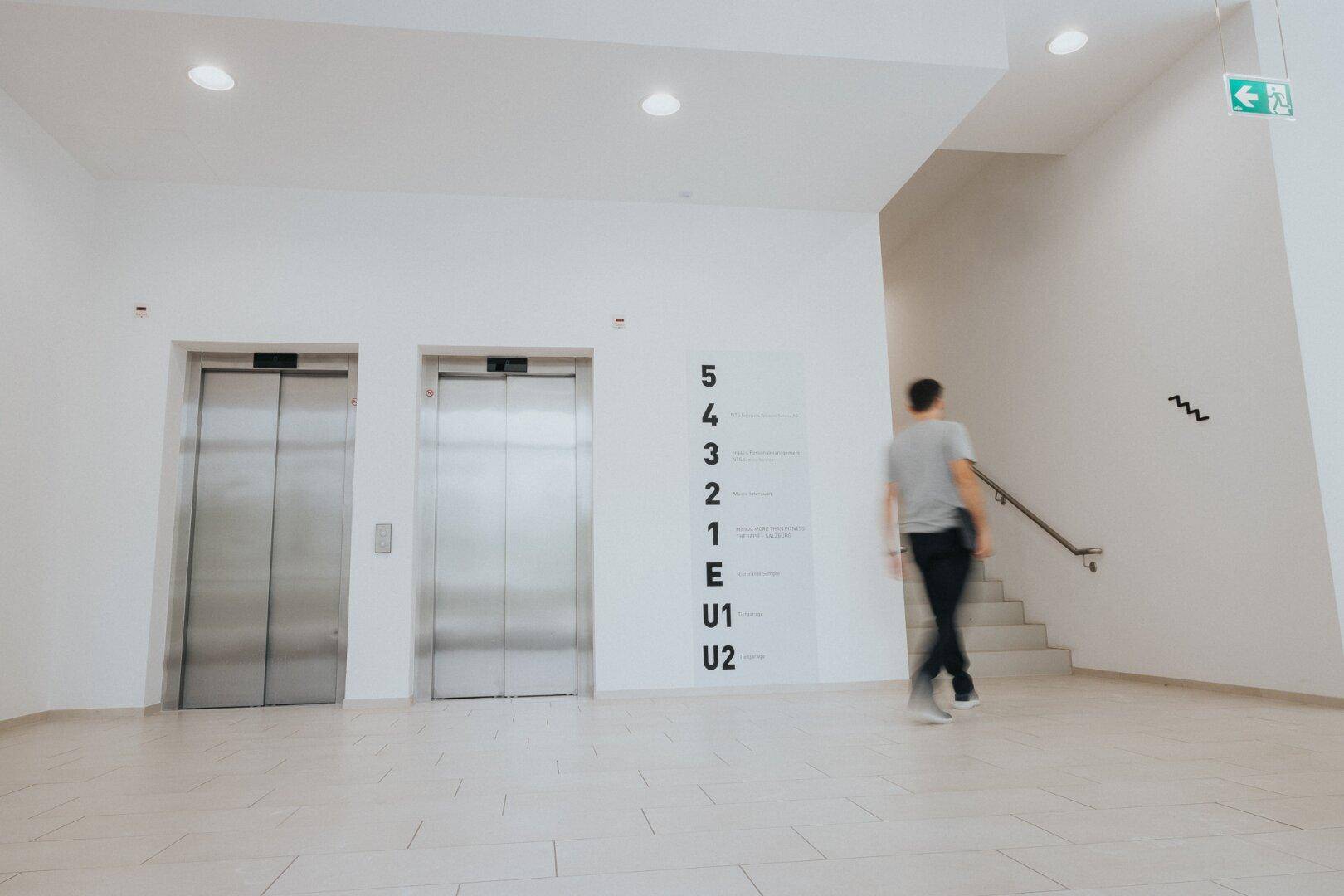 Heller Empfangsbereich mit zwei Aufzügen, einer Treppe und einem Personenleitsystem.