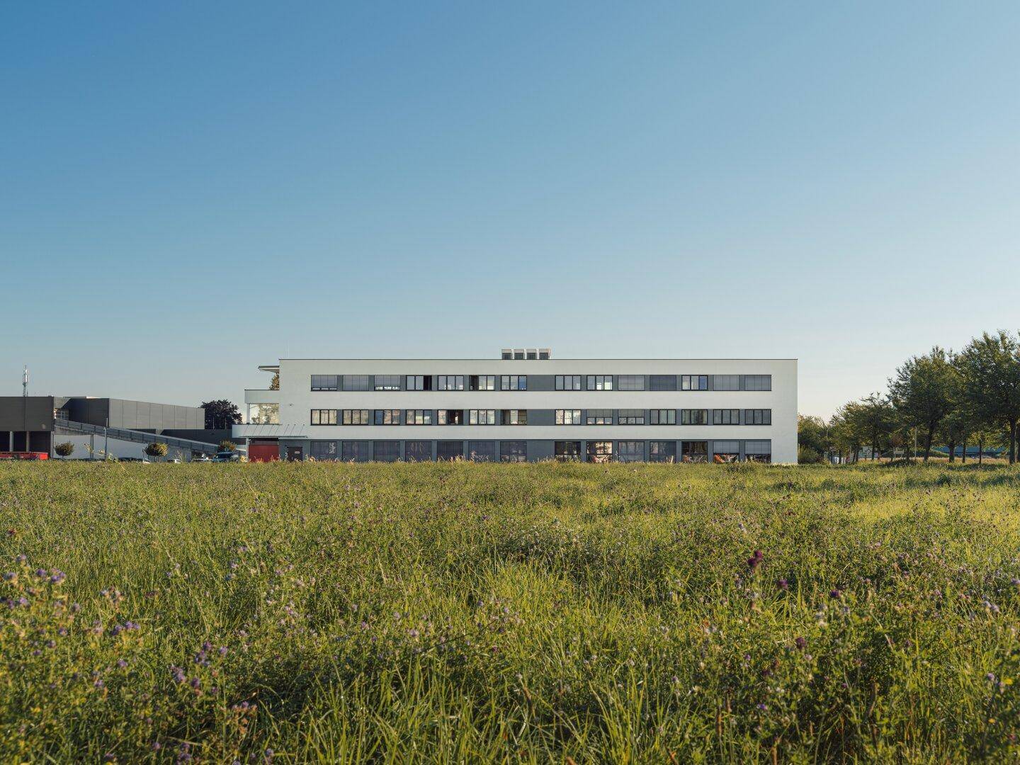 Modernes Bürogebäude in ländlicher Umgebung, umgeben von einer weiten Wiese unter blauem Himmel.