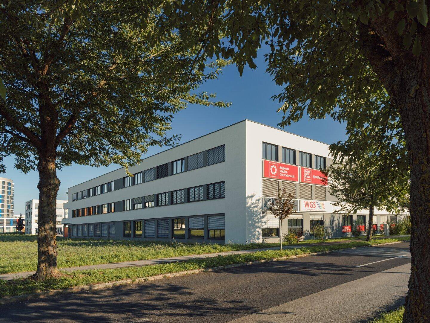 Modernes Bürogebäude mit weißer Fassade und großen Fenstern, umgeben von Bäumen und Grünflächen.