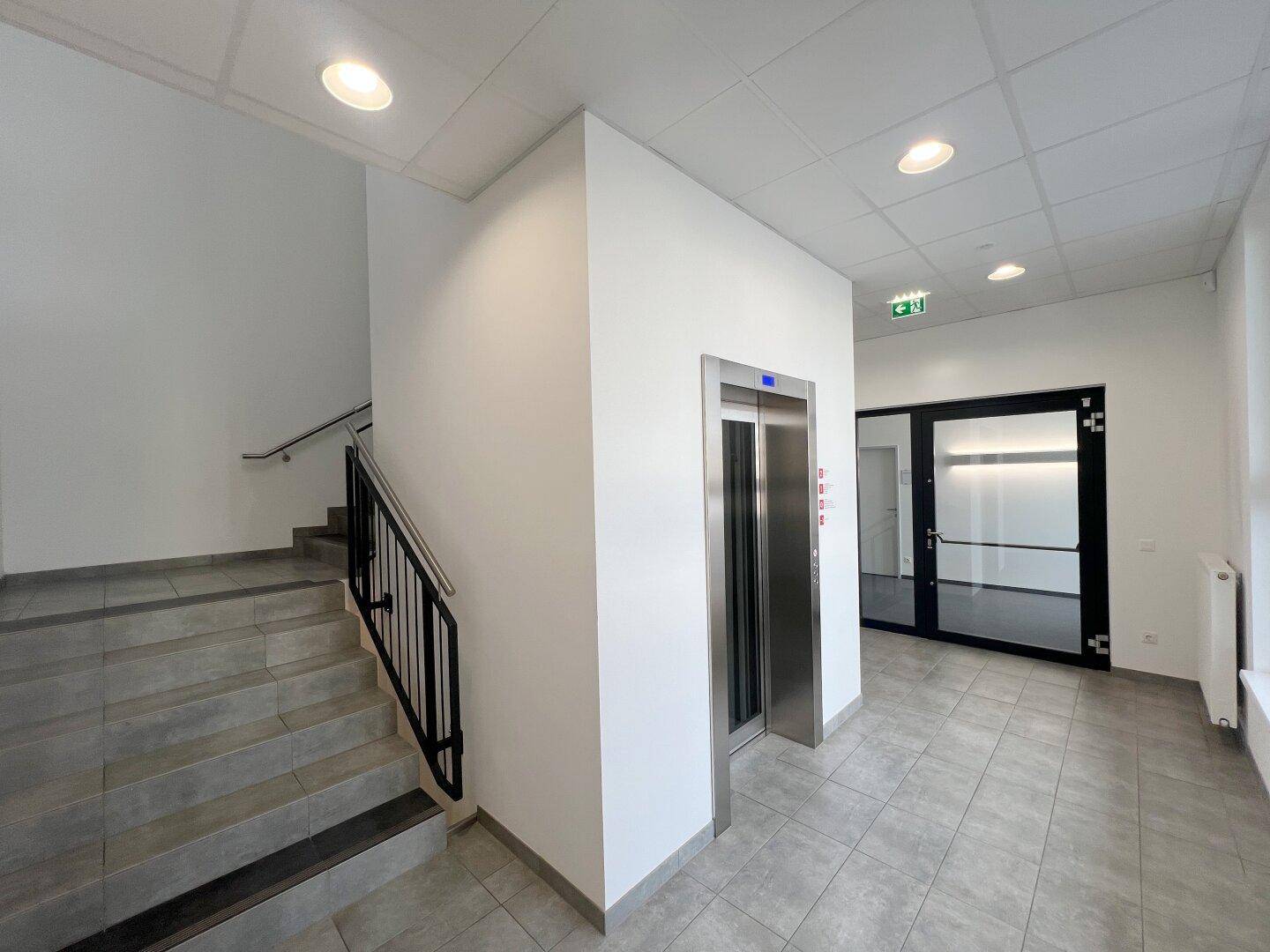 Moderner Eingangsbereich mit Treppe, Aufzug und Fliesenboden in hellem Grau, gut beleuchtet.