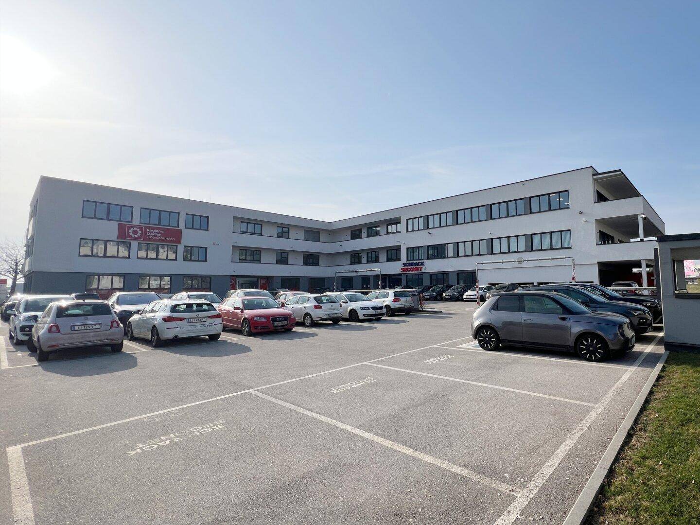 Großer Parkplatz vor einem modernen Bürogebäude unter klarem Himmel, mit vielen Fahrzeugen.