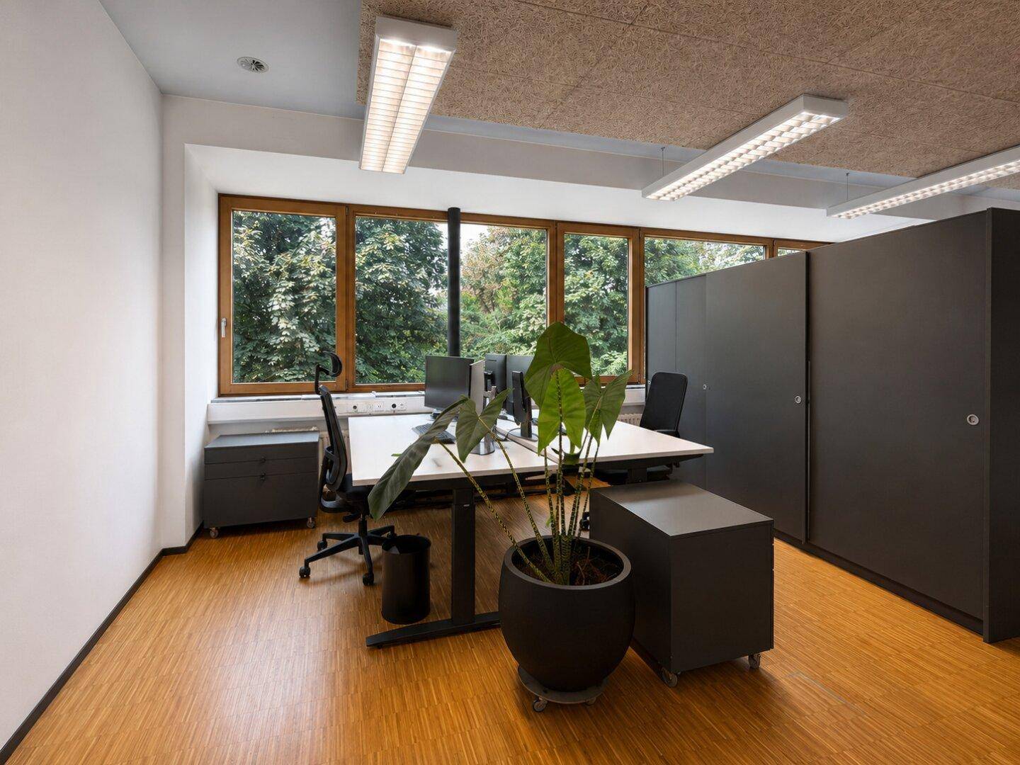 Einzelner Arbeitsplatz in einem hellen Büro mit Holzboden, großen Fenstern und Blick ins Grüne.
