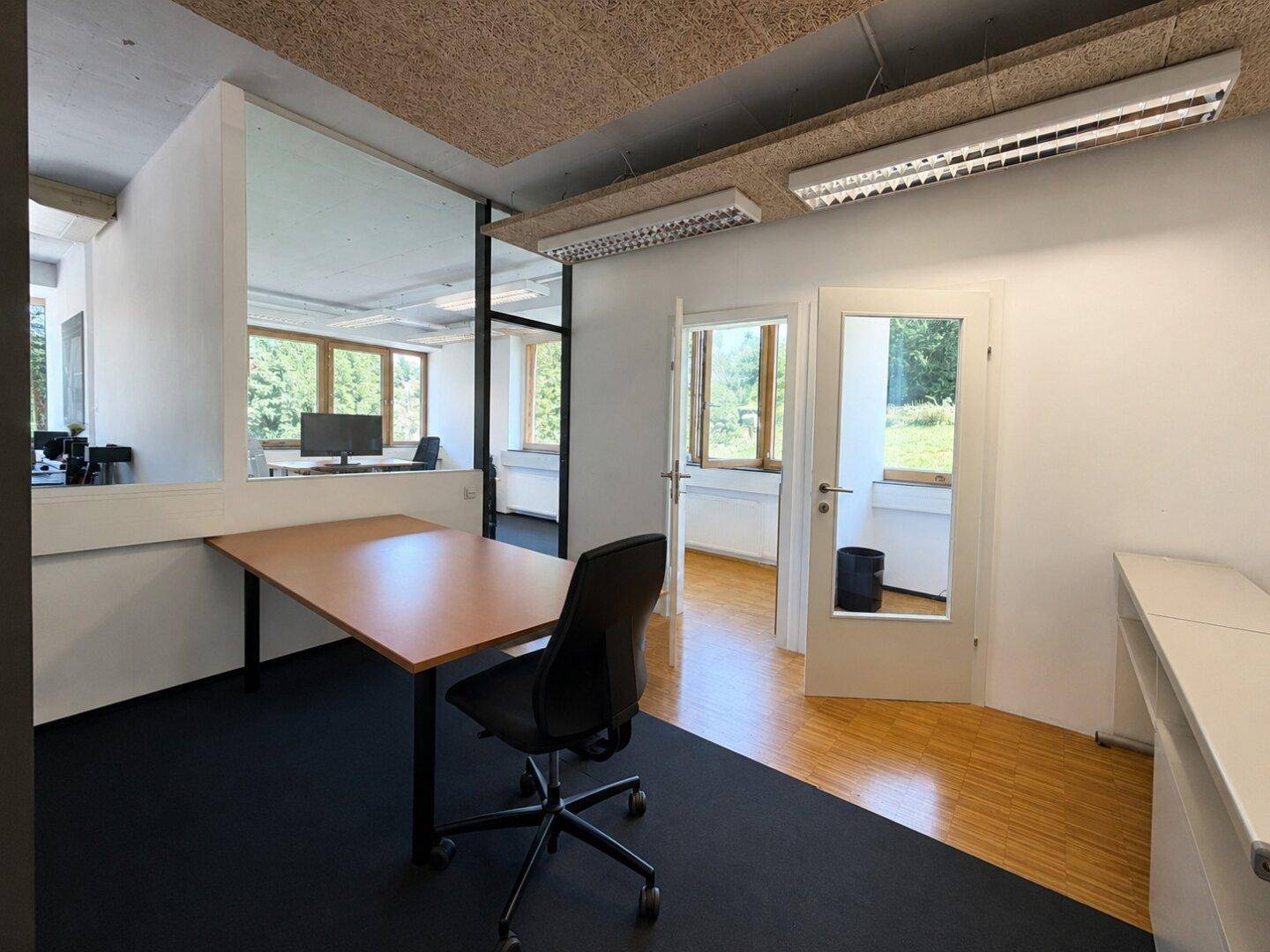 Büro mit Teppichboden und Holzbodenbereichen, Glaswand und Blick auf den Außenbereich.