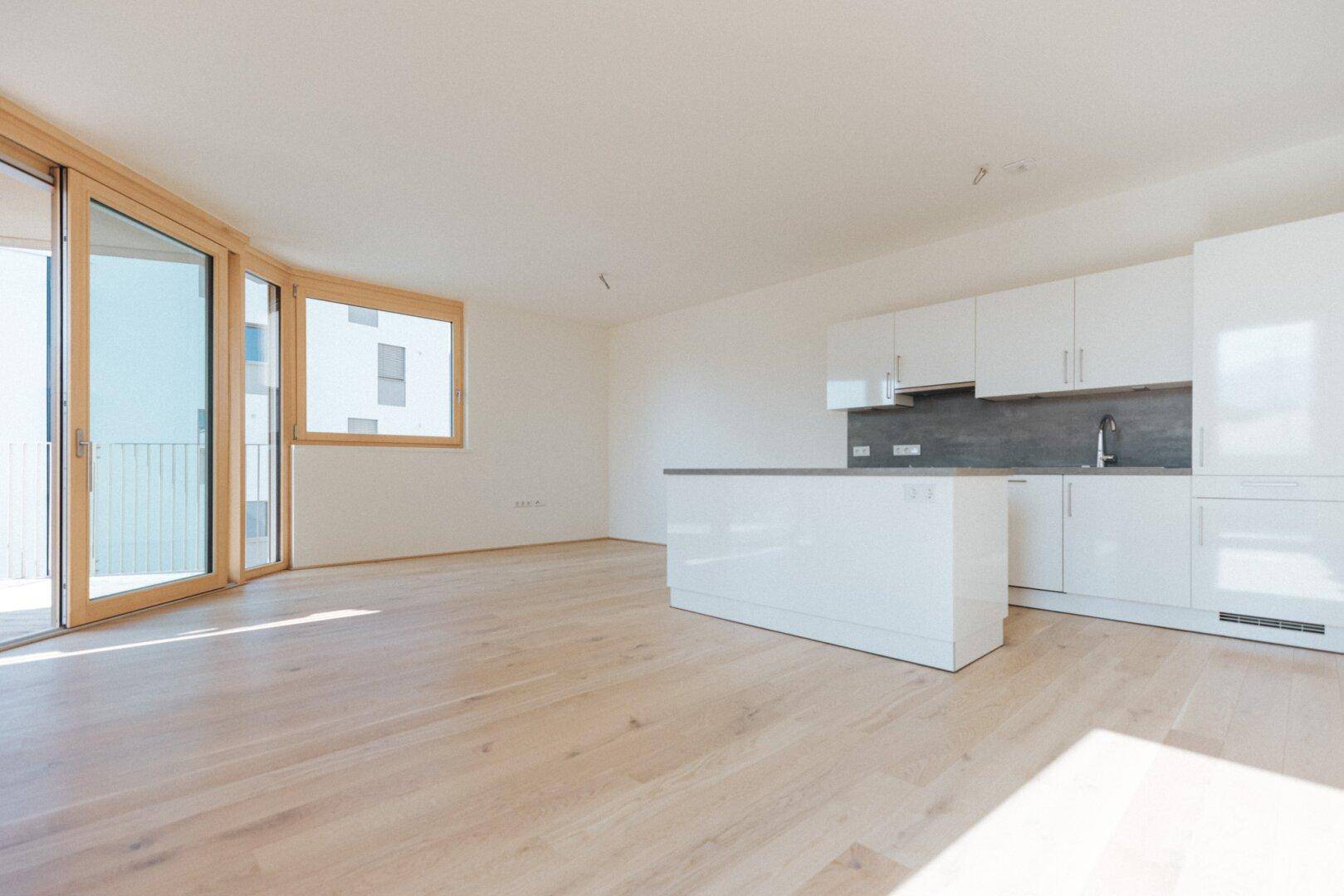 Offener Wohnbereich mit moderner weißer Küche, Holzboden und Zugang zum Balkon.