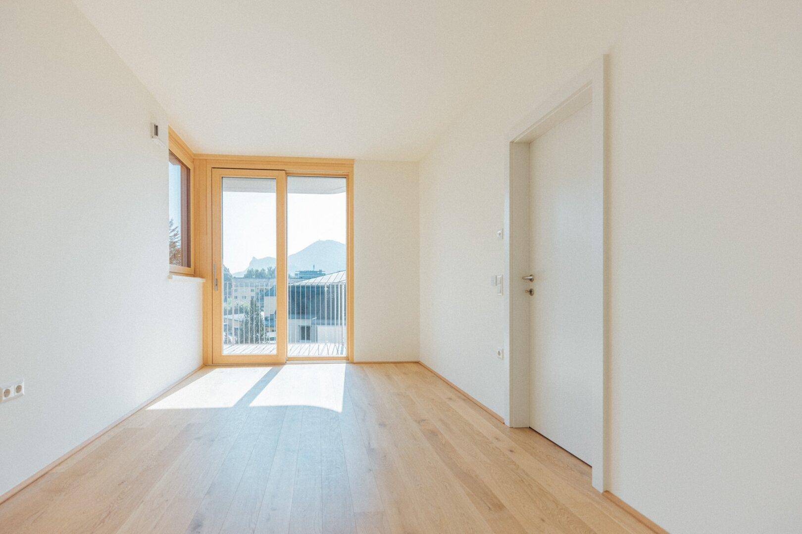 Heller Raum mit Holzboden, großen Fenstern und Zugang zu einem Balkon mit Bergblick.