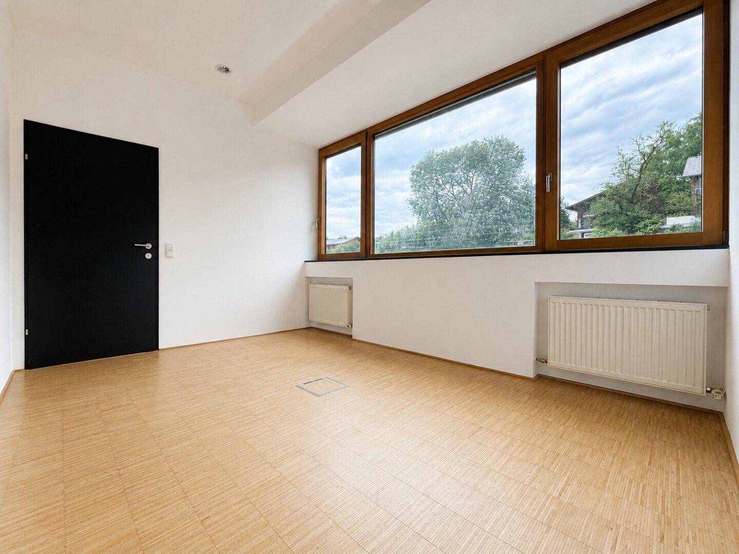 Heller Raum mit Holzboden, einer schwarzen Tür und großen Fenstern mit Ausblick.