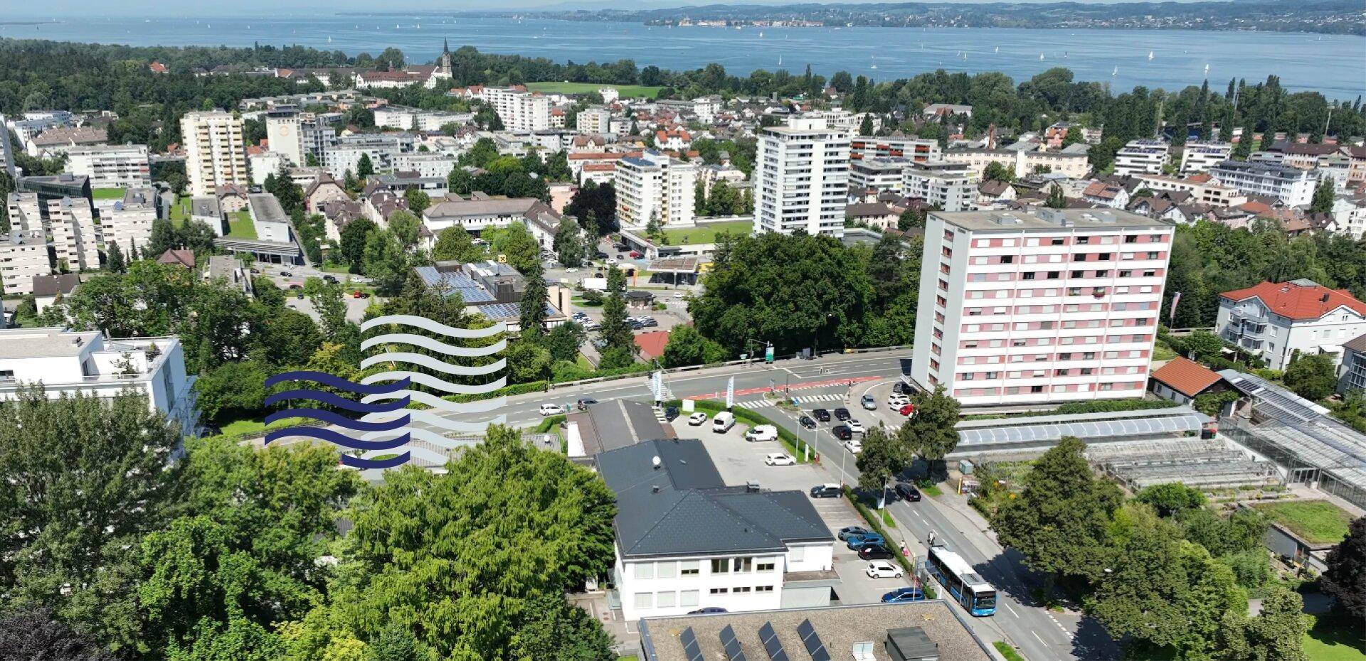 Luftaufnahme einer Stadtlandschaft mit zahlreichen Gebäuden, viel Grün und einem großen See im Hintergrund.