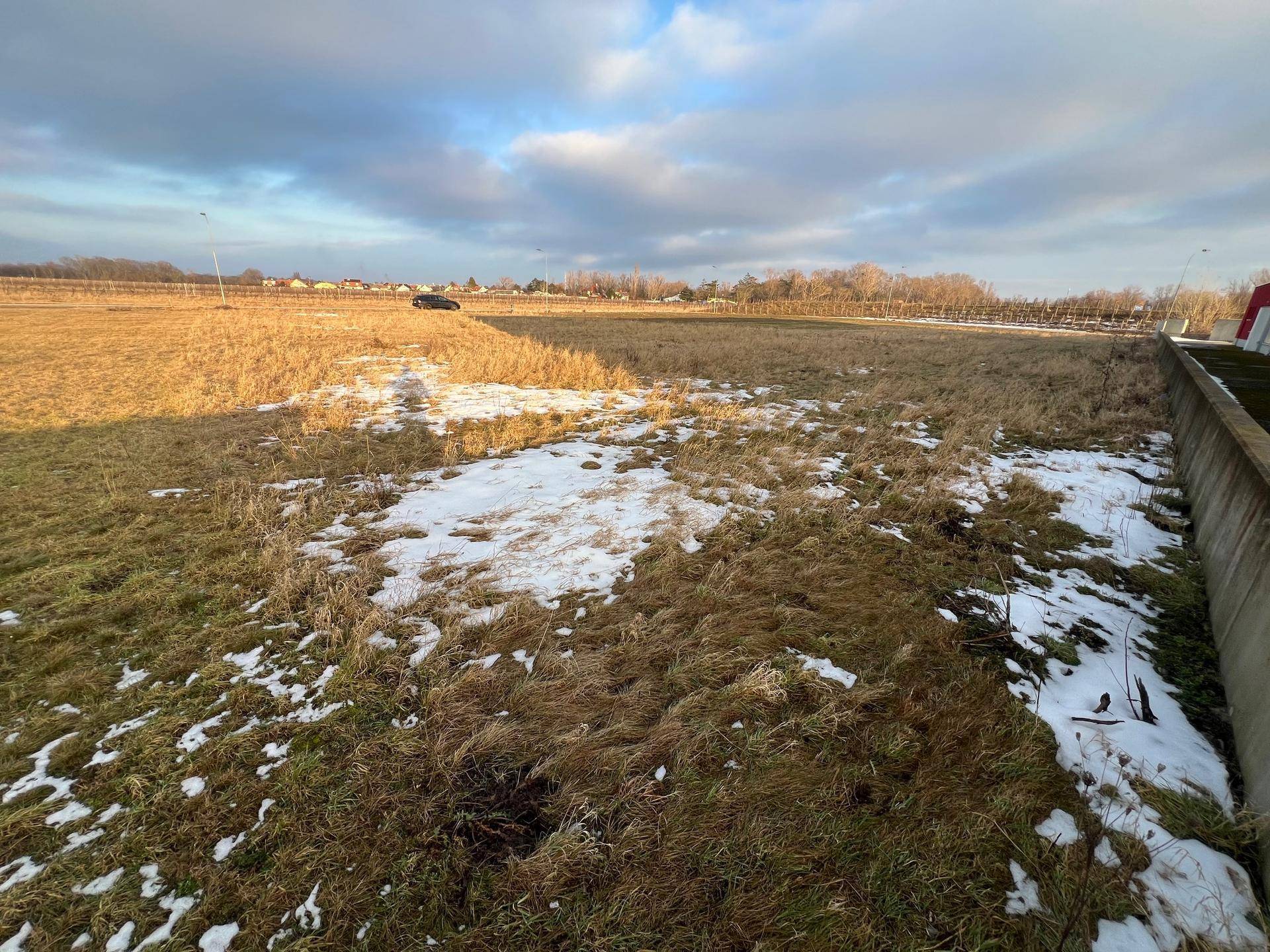 Weitläufiges, unbebautes Feld mit Schneeresten und einem Auto in der Ferne unter bewölktem Himmel.