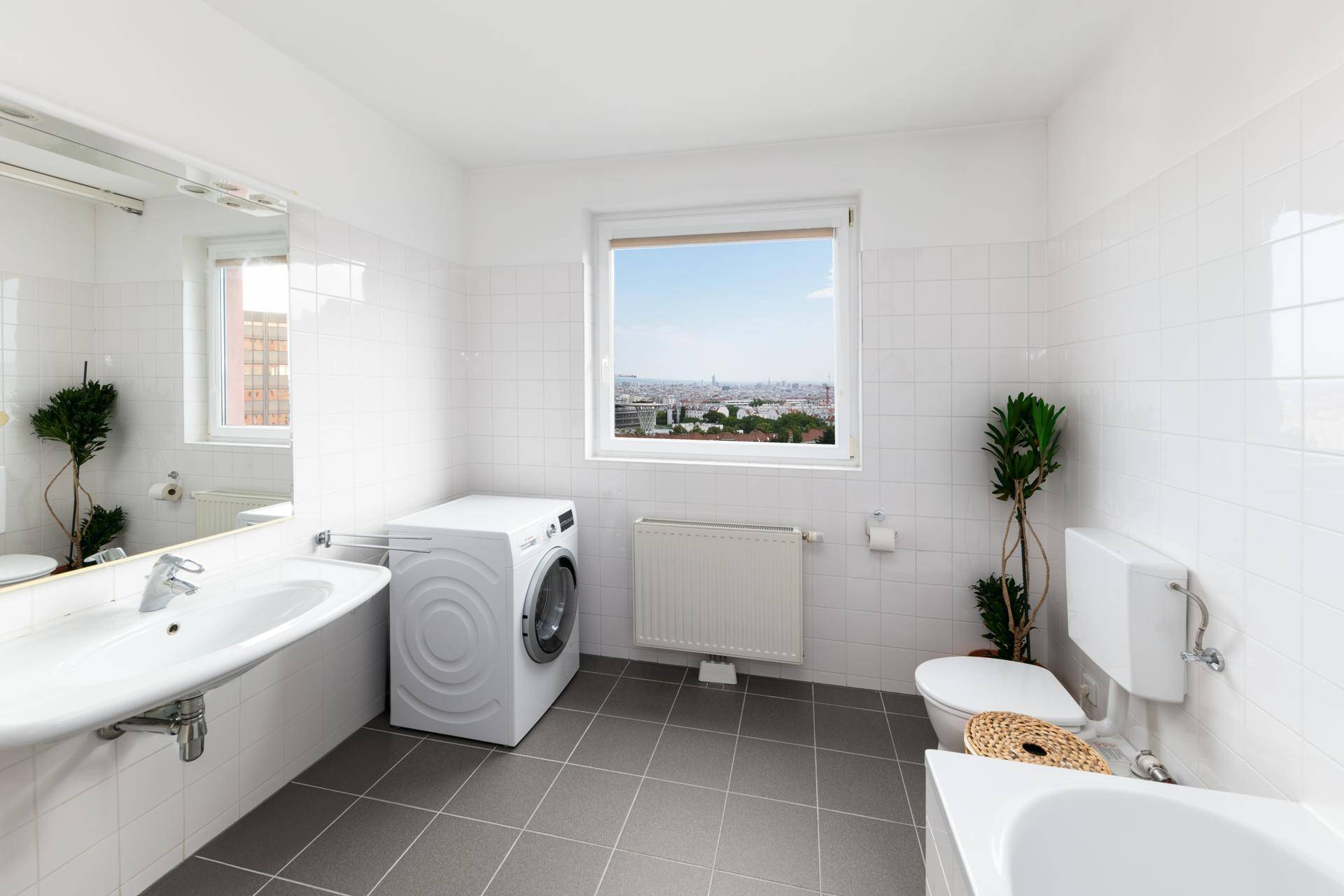 Badezimmer mit grauen Fliesen, Badewanne, WC und Fenster mit Stadtblick.
