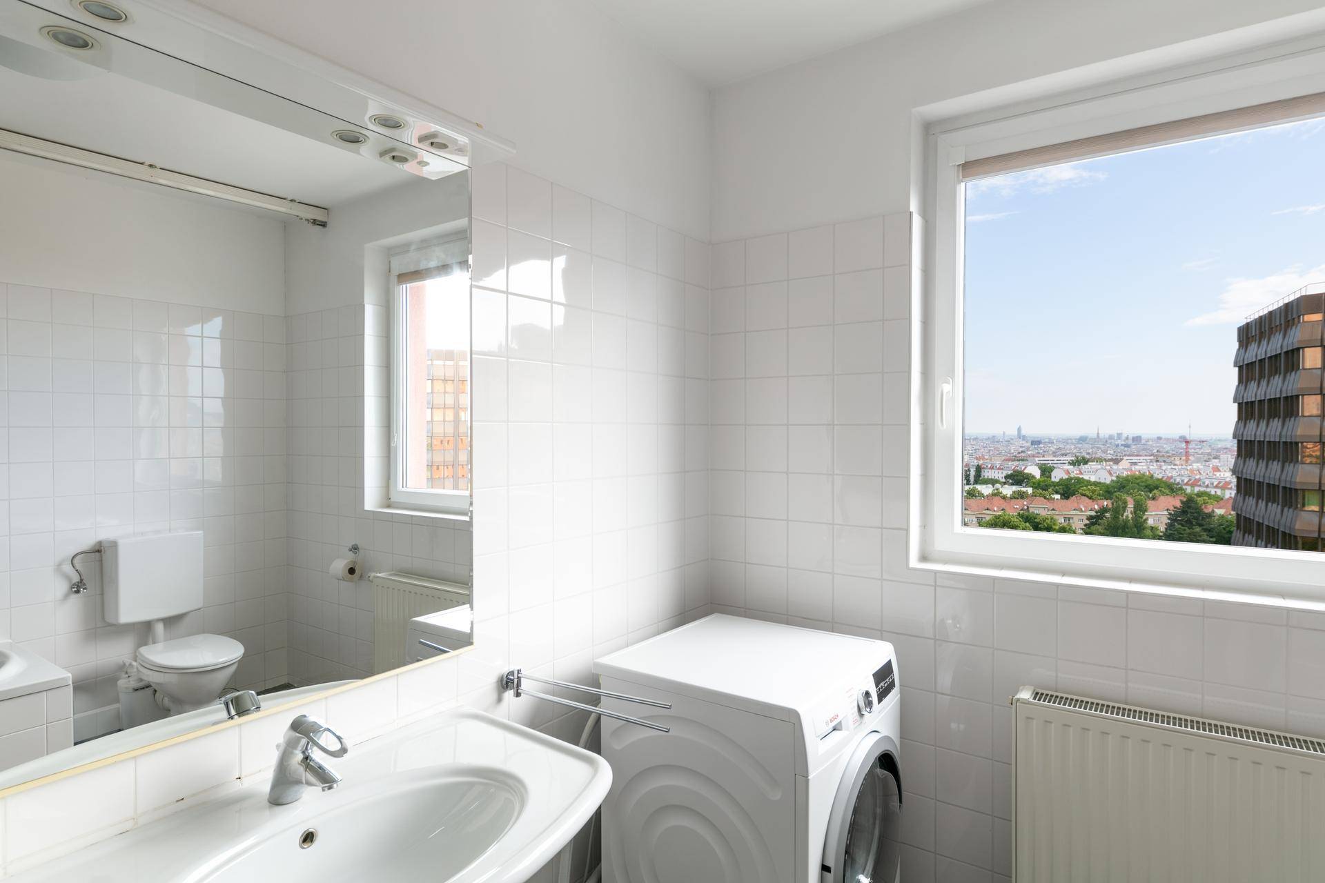 Helles Badezimmer mit weißen Fliesen, Waschmaschine und Fenster mit Stadtblick.