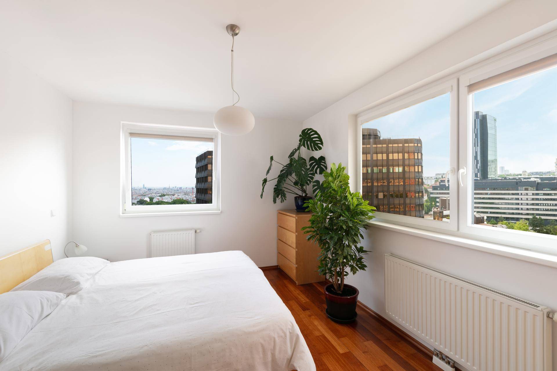 Helles Schlafzimmer mit Holzparkett, Doppelbett und Fenstern mit Stadtblick.