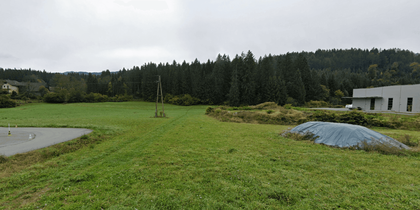 Weitläufige grüne Wiese mit dichtem Wald im Hintergrund und einem modernen Gebäude am rechten Bildrand.