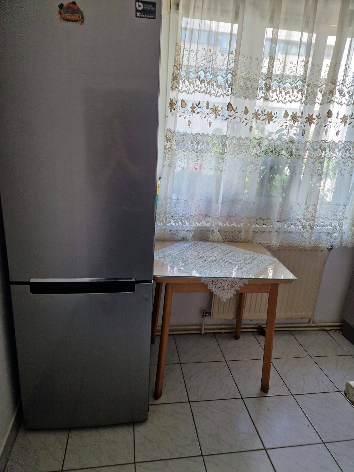 Küchenbereich mit einem modernen Kühlschrank, einem kleinen Esstisch und einem Fenster mit Spitzenvorhängen.