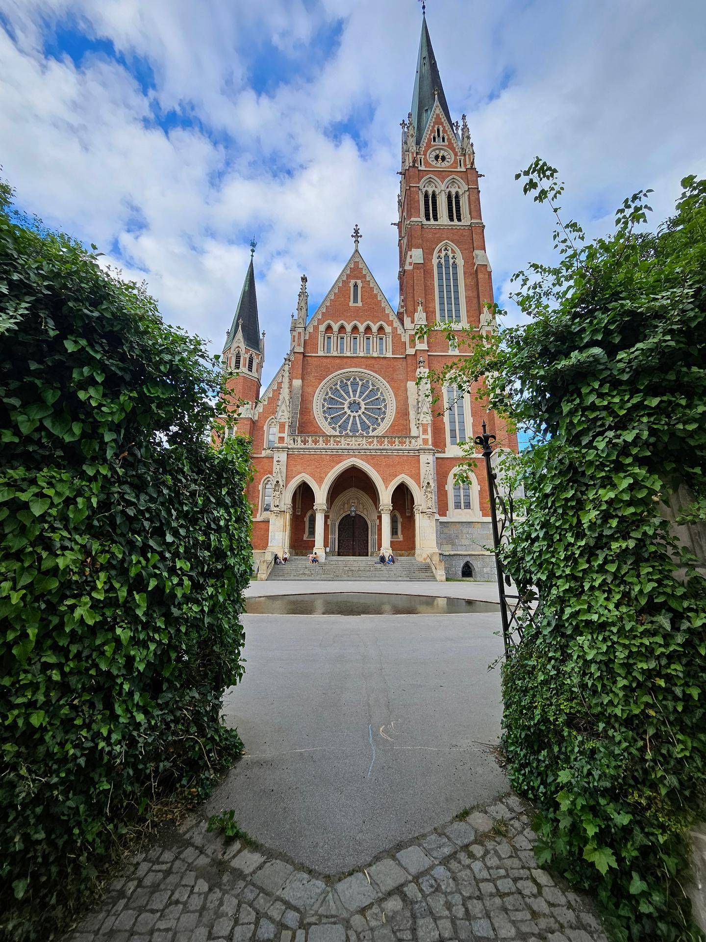 Beeindruckende gotische Kirche mit roten Ziegeln und hohen Türmen, umgeben von Grün.