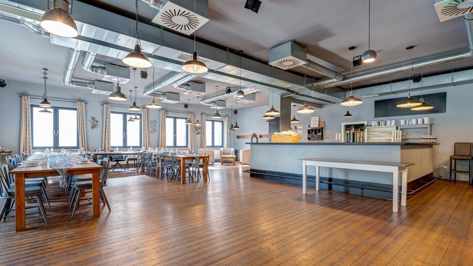 Geräumiger Veranstaltungsraum mit Holzboden, vielen Tischen und einer modernen Bar im Hintergrund.